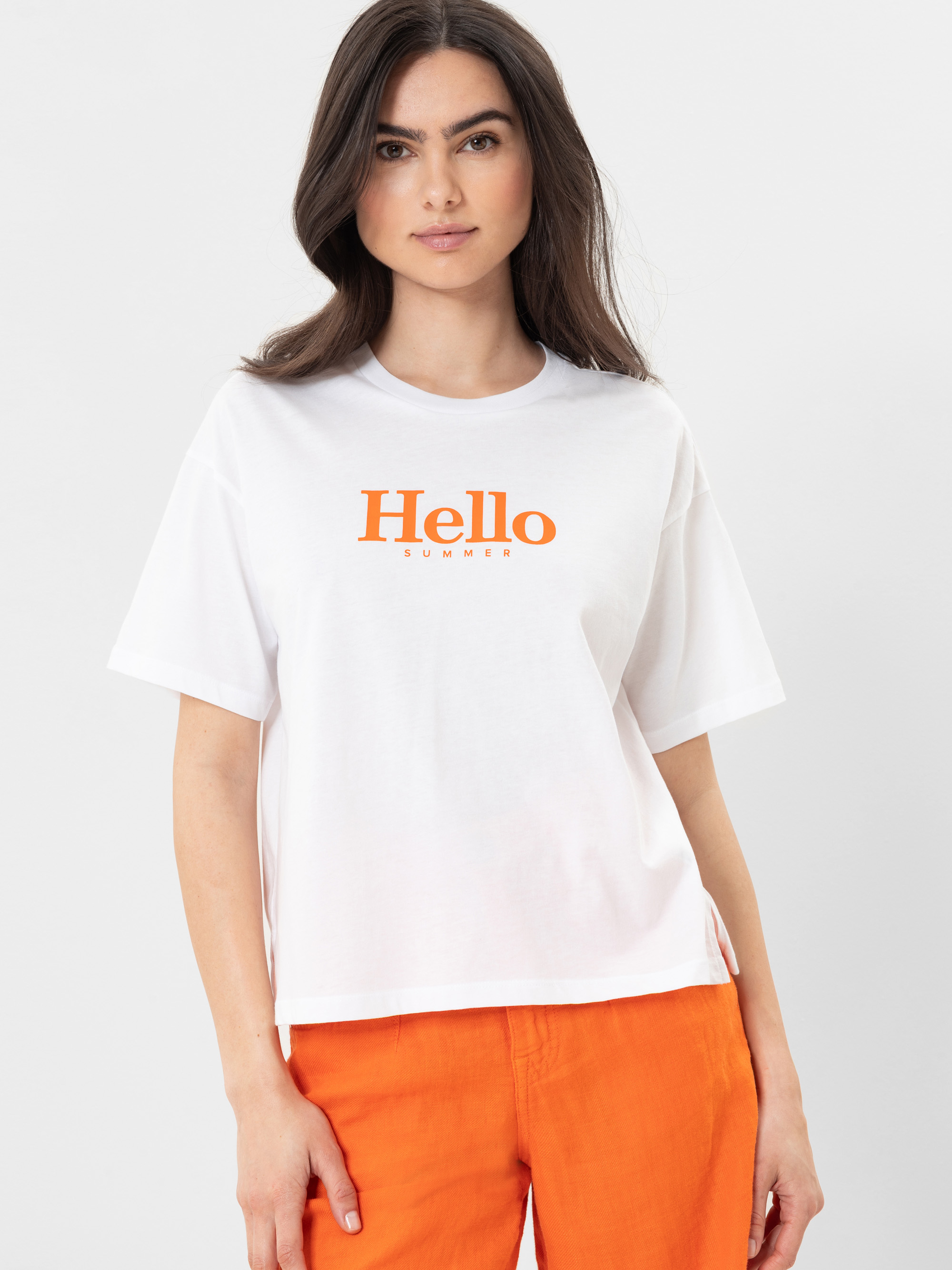 Eine Frau mit langen braunen Haaren, die ein weißes T-Shirt mit der Aufschrift Hello Summer in orangefarbener Schrift und eine orangefarbene Hose trägt, steht vor einem schlichten hellen Hintergrund.