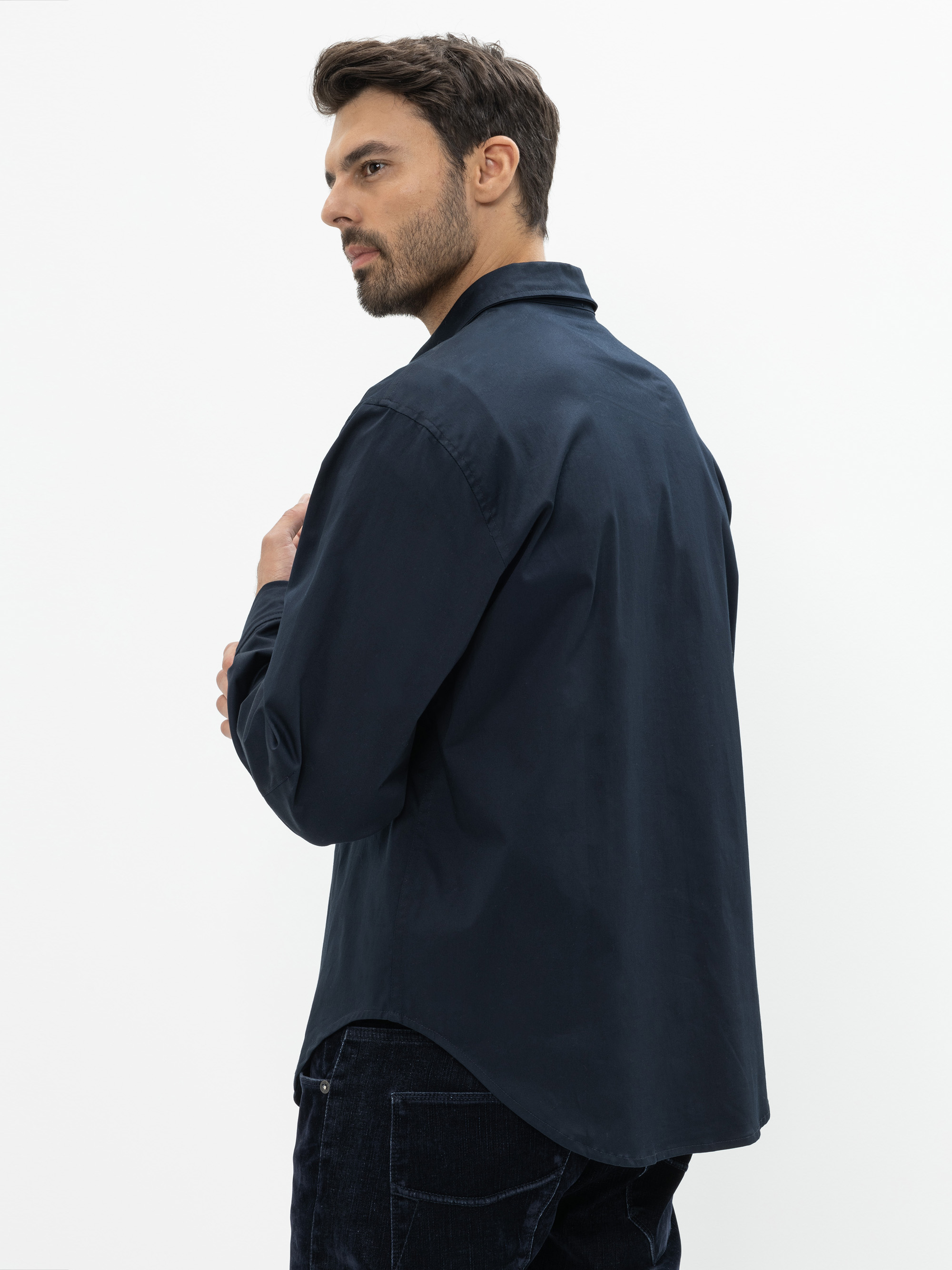 Ein Mann mit kurzen braunen Haaren und einem Bart trägt ein dunkles, marineblaues, langärmeliges Hemd und eine dunkle Hose. Er steht seitlich und schaut über seine Schulter vor einem schlichten weißen Hintergrund.