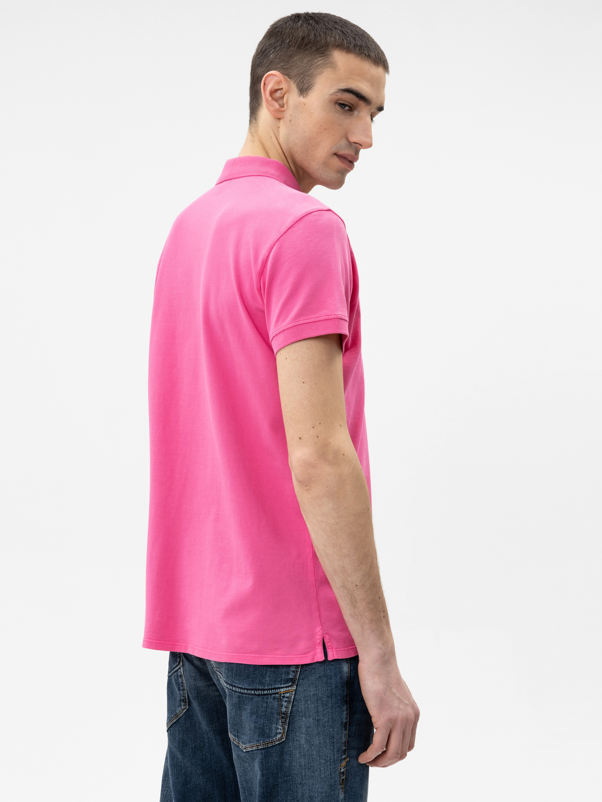 Eine Person mit kurzen braunen Haaren, die ein hellrosa Polohemd und blaue Jeans trägt, steht leicht zur Seite gedreht und blickt über ihre Schulter auf einen schlichten weißen Hintergrund.