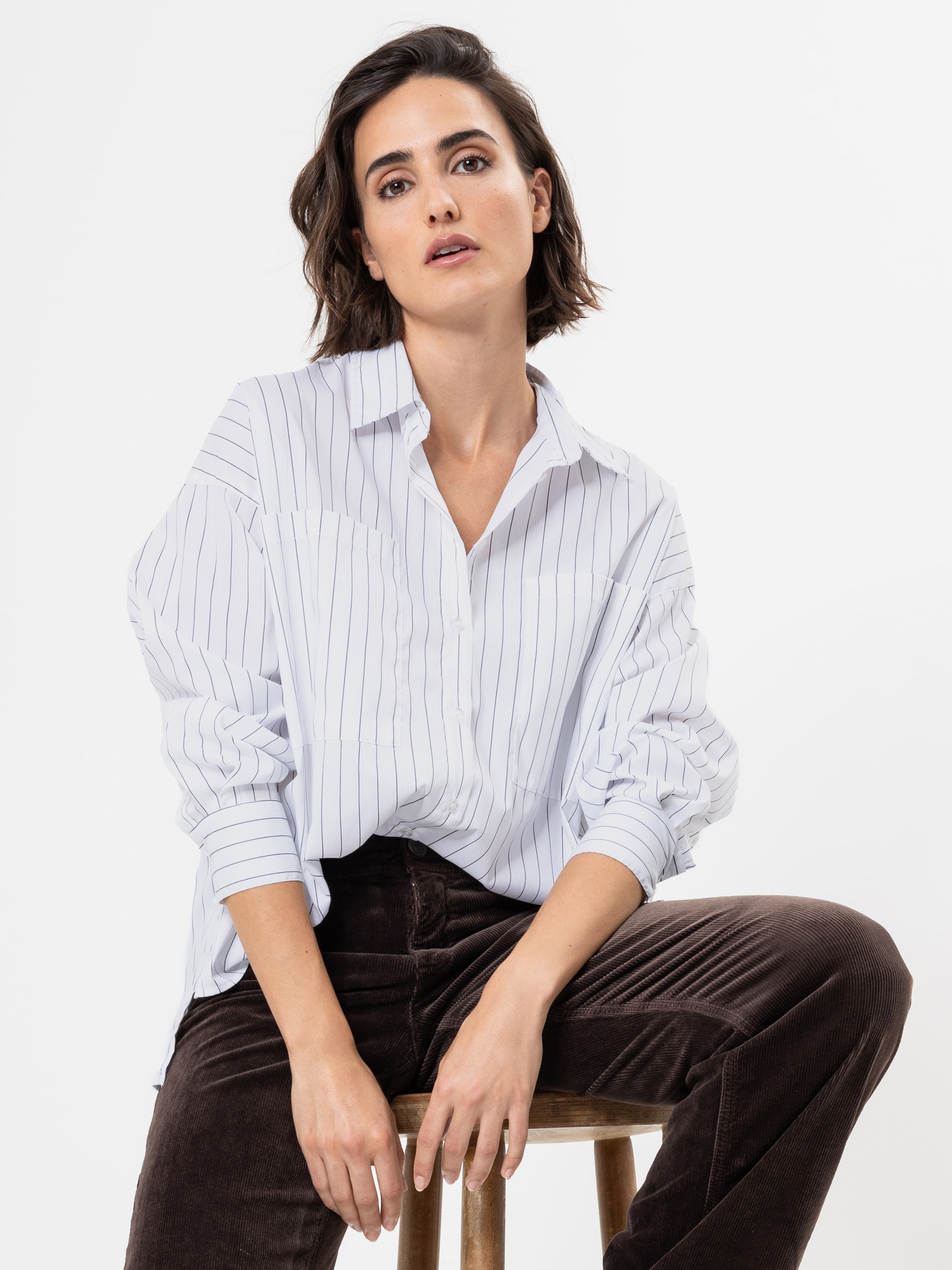 Eine Frau mit kurzen braunen Haaren sitzt auf einem Holzschemel vor einem weißen Hintergrund. Sie trägt ein weißes Hemd mit dünnen vertikalen Streifen und eine braune Hose. Sie nimmt eine entspannte Haltung ein, wobei ihre Hände auf den Knien ruhen.