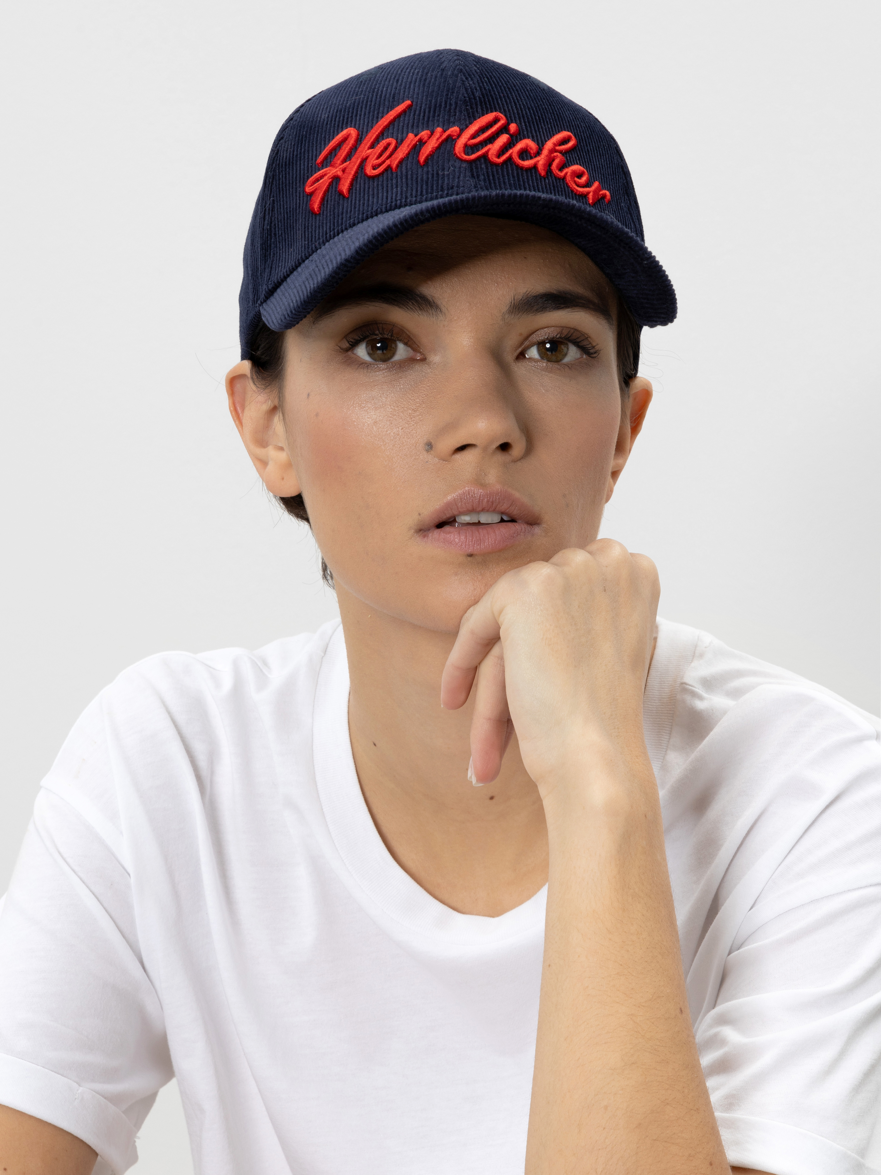 Eine Person, die eine marineblaue Mütze mit dem Wort Herrlicher in roter Schrift und ein schlichtes weißes T-Shirt trägt, stützt ihr Kinn auf die Hand und blickt leicht nach oben.