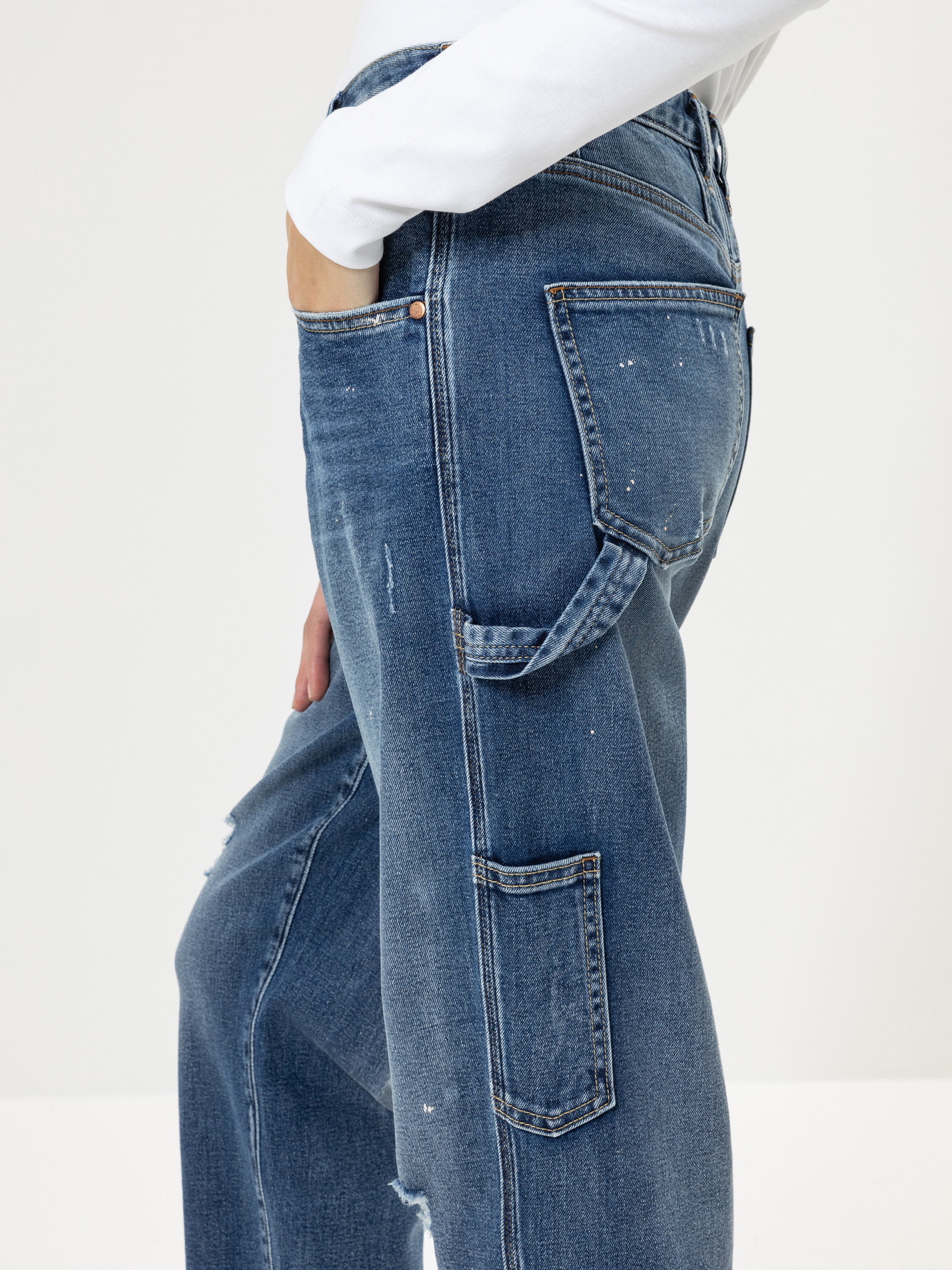Eine Person, die eine blaue Jeans mit mehreren Taschen und ein weißes langärmeliges Hemd trägt, wird von der Hüfte abwärts in einer Seitenansicht vor einem unifarbenen Hintergrund gezeigt.