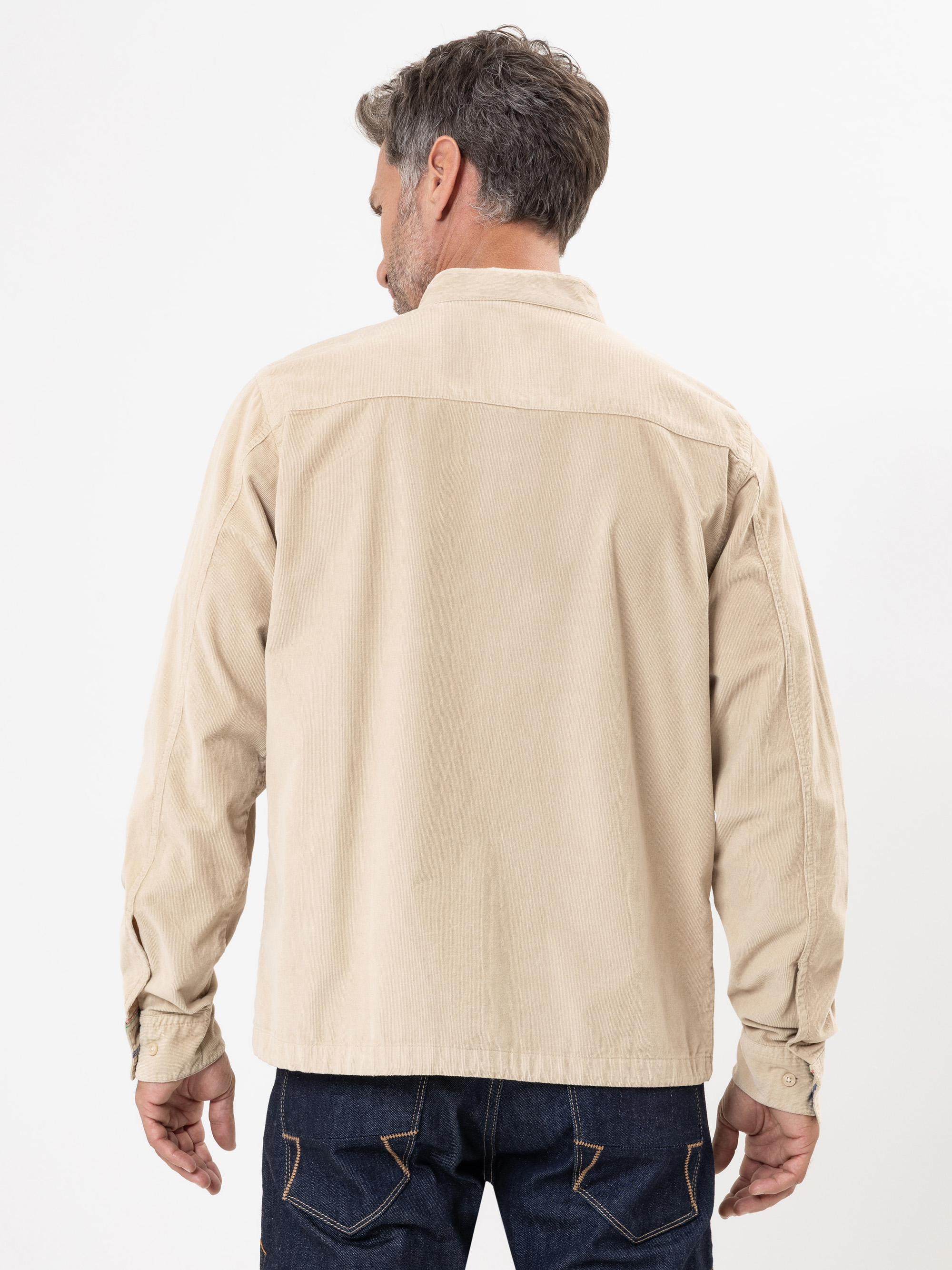 Ein Mann mit kurzen, dunklen Haaren wird von hinten gezeigt, der eine hellbeige Kordjacke und dunkelblaue Jeans vor einem schlichten weißen Hintergrund trägt.