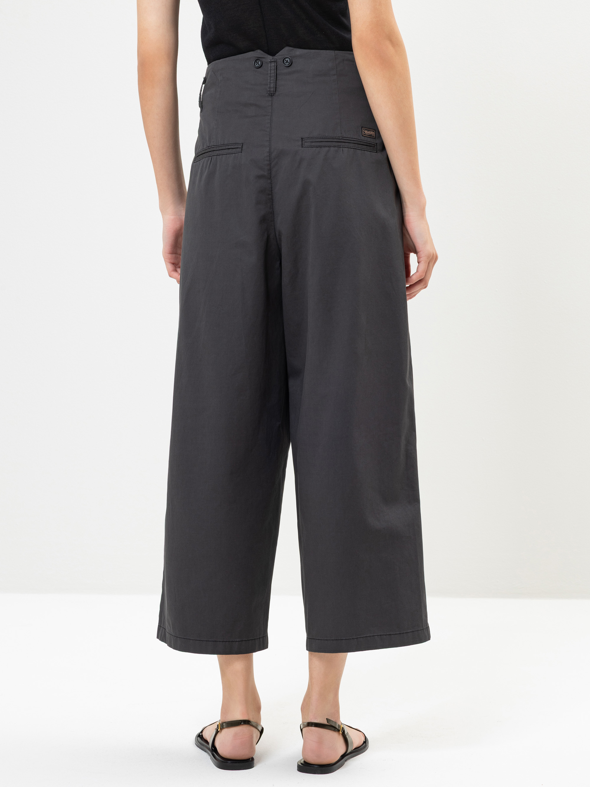 Eine Person, die eine weite, hoch taillierte, dunkelgraue Hose und schwarze Sandalen trägt, steht mit dem Rücken zur Kamera vor einem schlichten weißen Hintergrund.