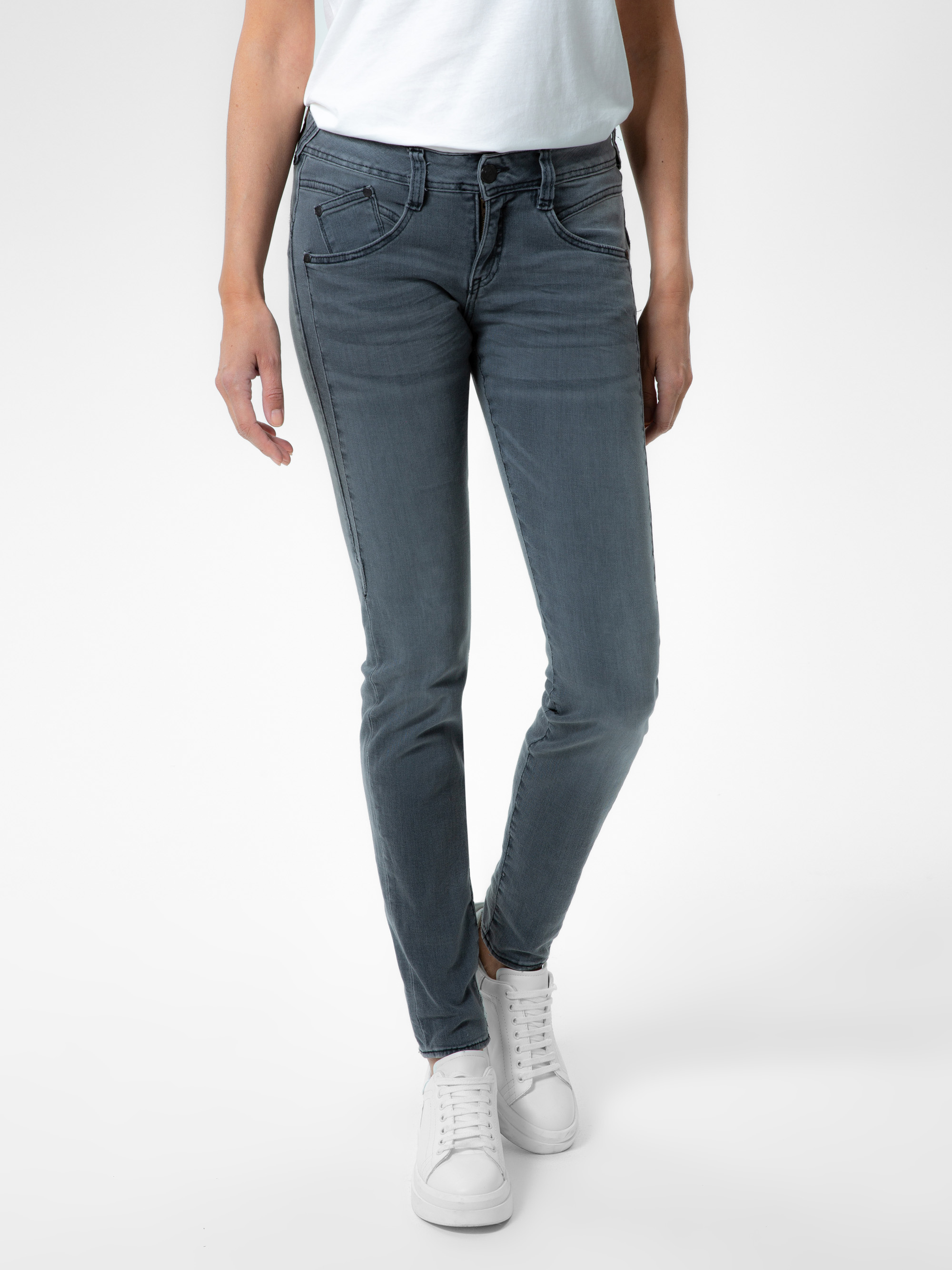 Eine Person, die ein weißes T-Shirt, graue Skinny Jeans und weiße Turnschuhe trägt, steht vor einem schlichten weißen Hintergrund. Das Bild konzentriert sich auf die untere Hälfte des Körpers.