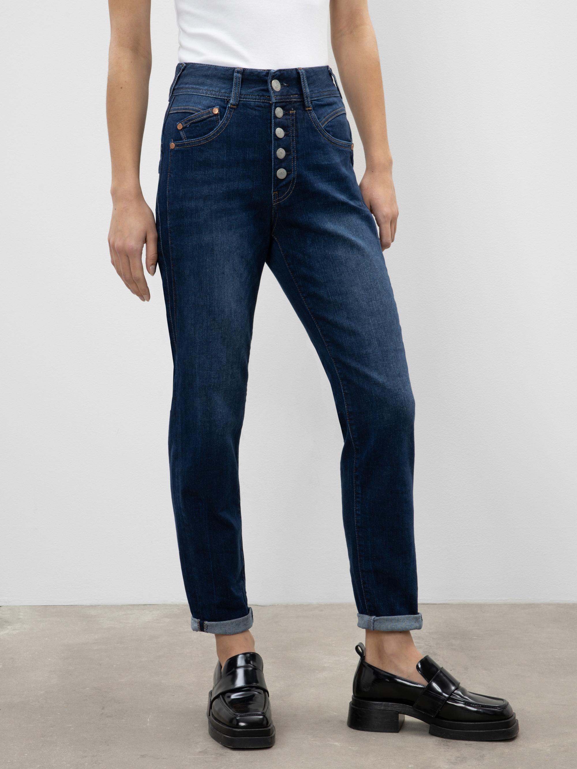 Eine Person, die dunkelblaue, hochgekrempelte Jeans mit auffälligen Knöpfen, ein weißes, ärmelloses Oberteil und schwarze Slipper trägt, steht vor einem schlichten, hellen Hintergrund.