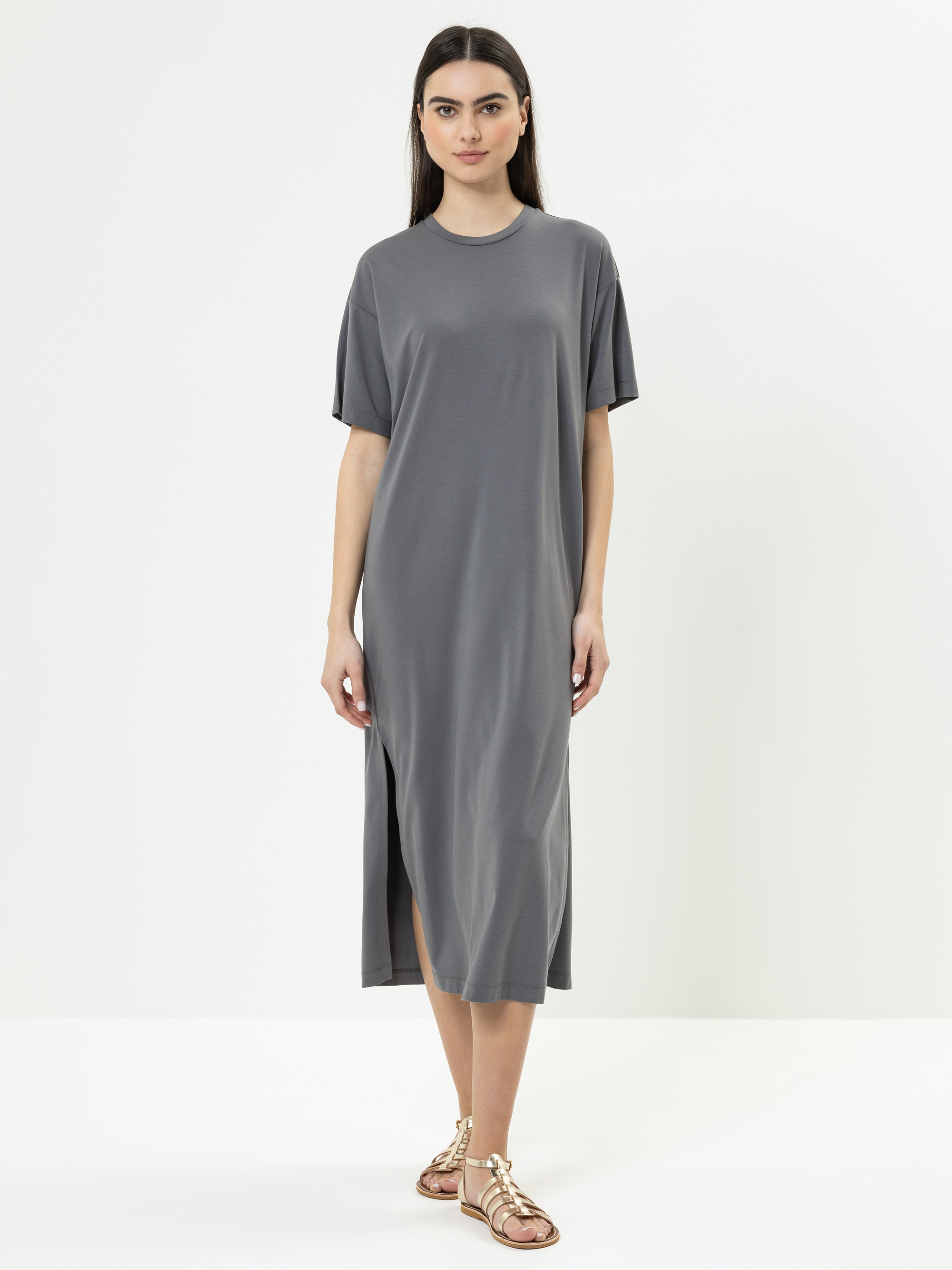 Eine Frau steht vor einem schlichten weißen Hintergrund und trägt ein locker sitzendes, kurzärmeliges, graues Midikleid mit Seitenschlitzen und flache Riemchensandalen. Sie hat glattes dunkles Haar und einen neutralen Gesichtsausdruck.