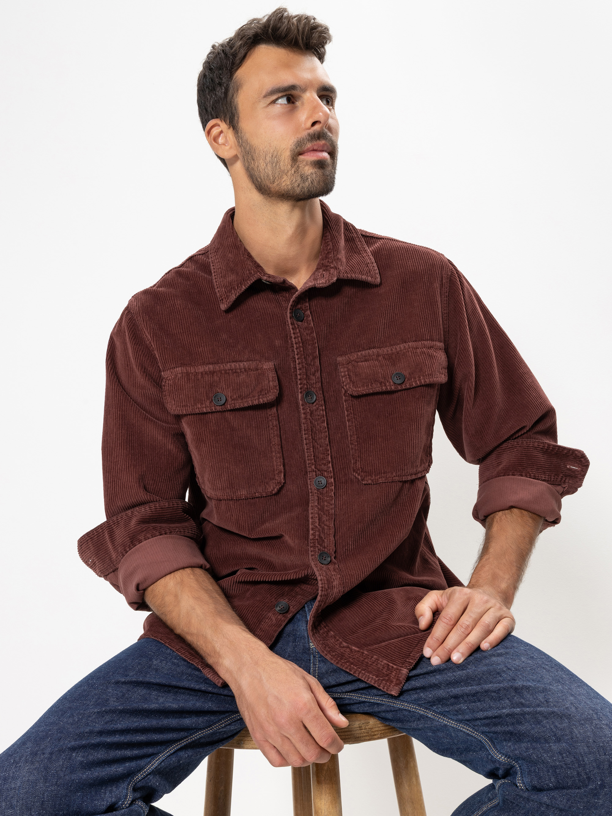 Ein Mann mit kurzen dunklen Haaren und einem Bart sitzt auf einem Hocker vor einem weißen Hintergrund. Er trägt ein braunes Cordhemd mit gekrempelten Ärmeln und blaue Jeans. Er blickt leicht zur Seite und hat einen neutralen Blick.