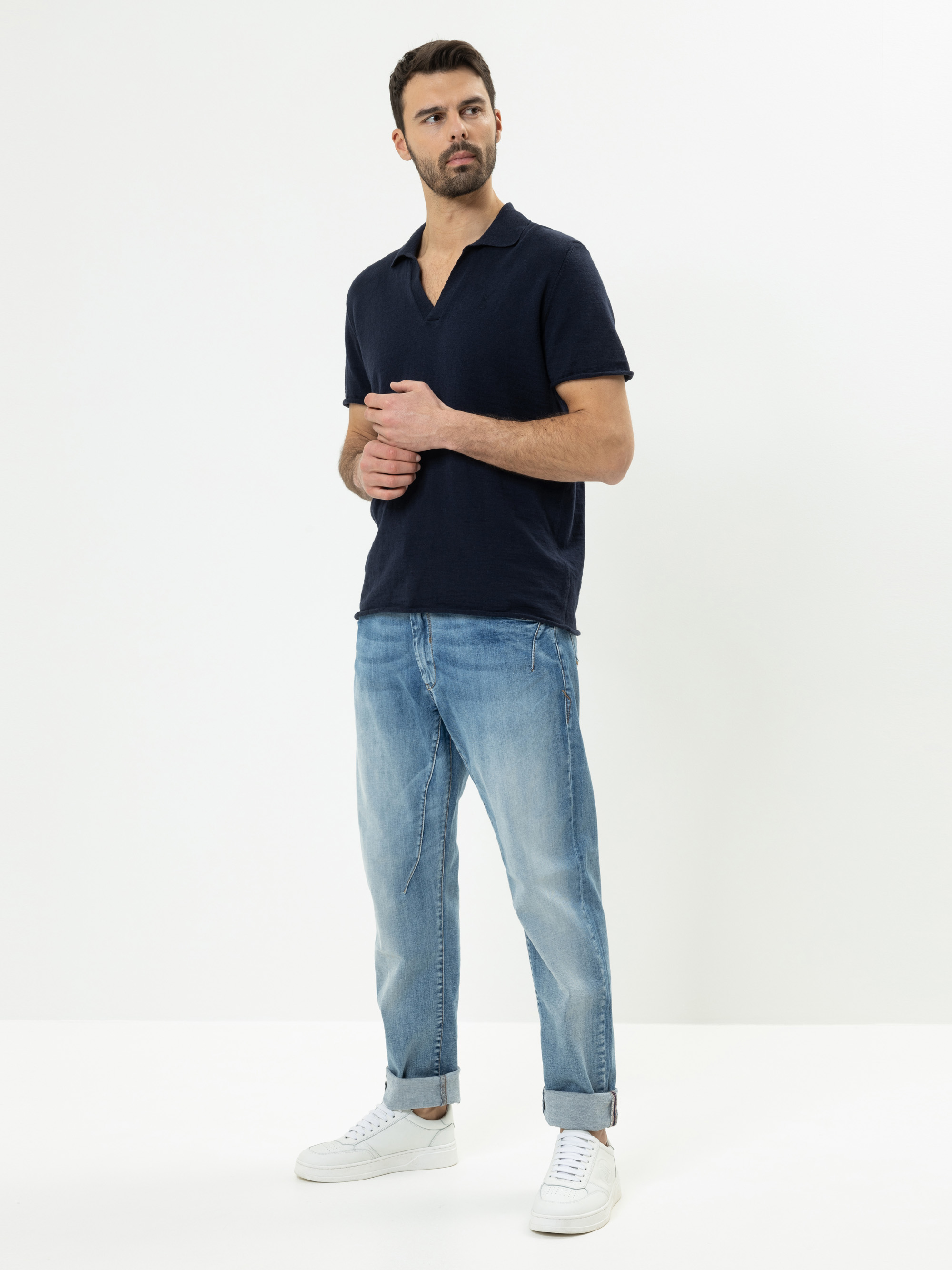 Ein Mann steht in einem minimalistischen Studio und trägt ein marineblaues Poloshirt, hellblaue Jeans mit Manschetten und weiße Turnschuhe. Er blickt zur Seite, die Hände locker vor sich verschränkt, vor einem schlichten weißen Hintergrund.