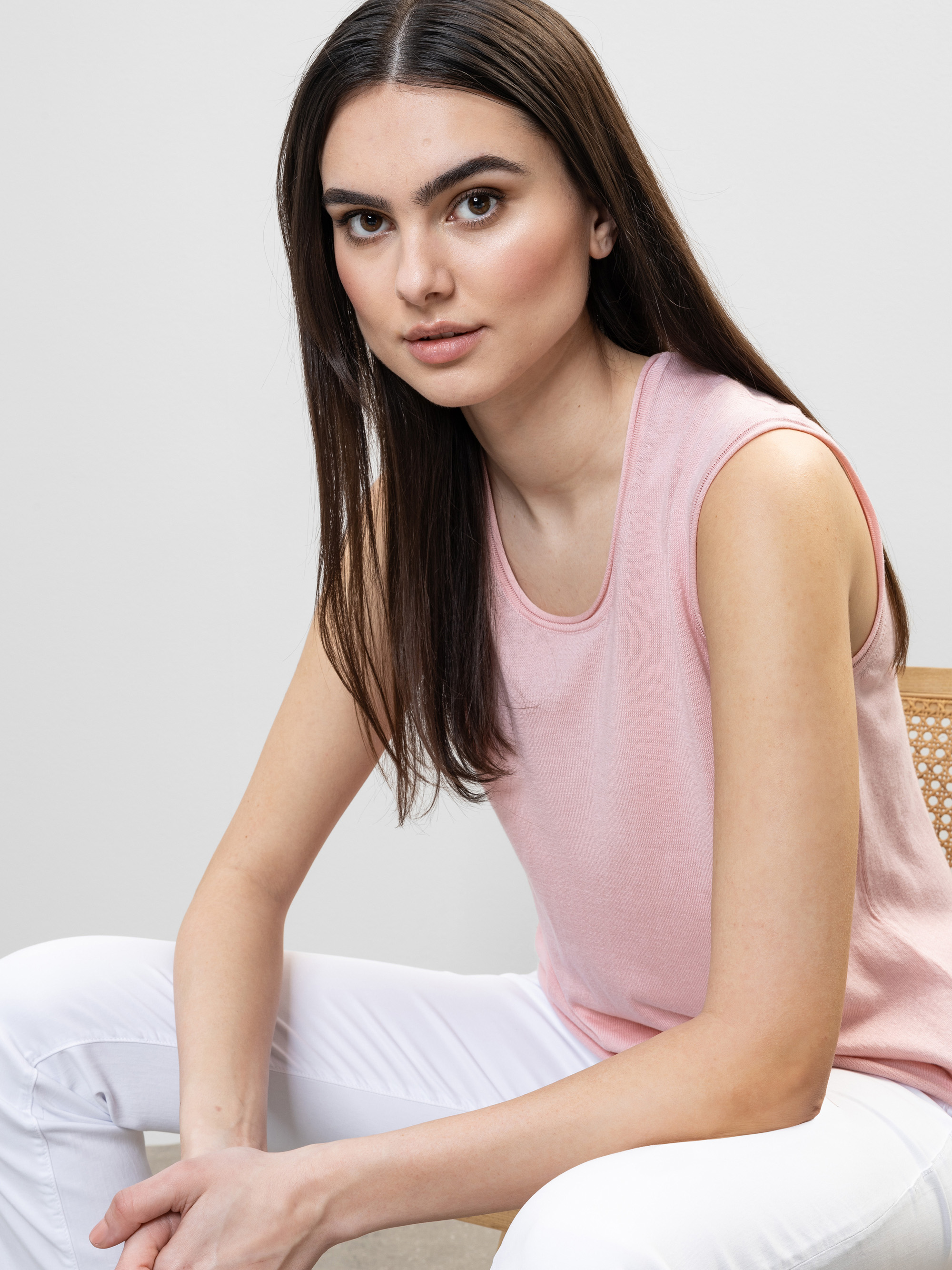 Eine Frau mit langen braunen Haaren sitzt auf einem geflochtenen Stuhl, trägt ein ärmelloses rosafarbenes Oberteil und eine weiße Hose und blickt mit neutralem Gesichtsausdruck direkt in die Kamera vor einem schlichten Hintergrund.