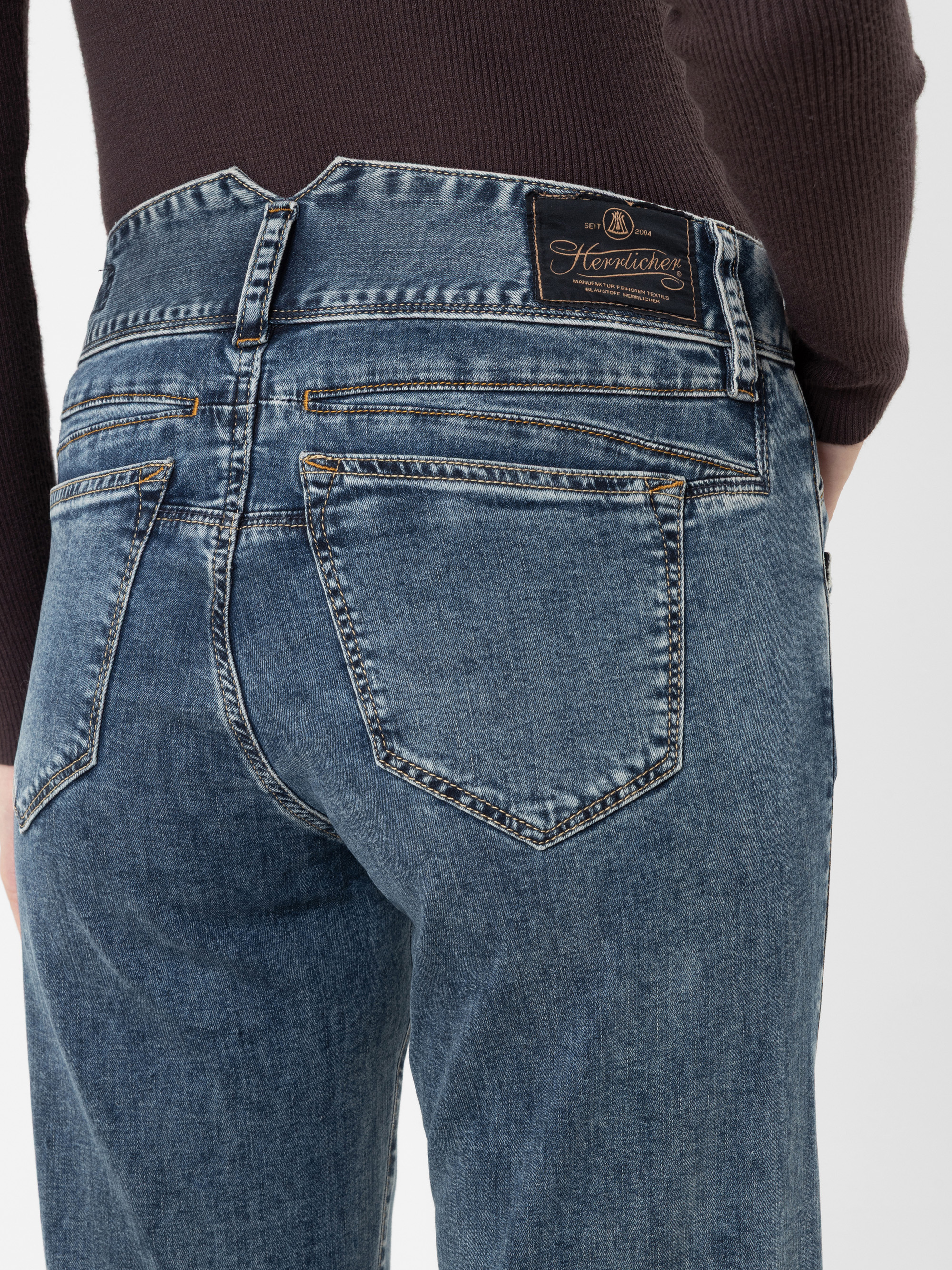 Eine Rückansicht einer Person, die eine blaue „Herrlicher“-Jeans mit einem markanten, hoch geschnittenen, V-förmigen Bund und einem sichtbaren schwarzen Markenetikett auf der Rückseite trägt, kombiniert mit einem dunkelbraunen Langarm-Oberteil vor