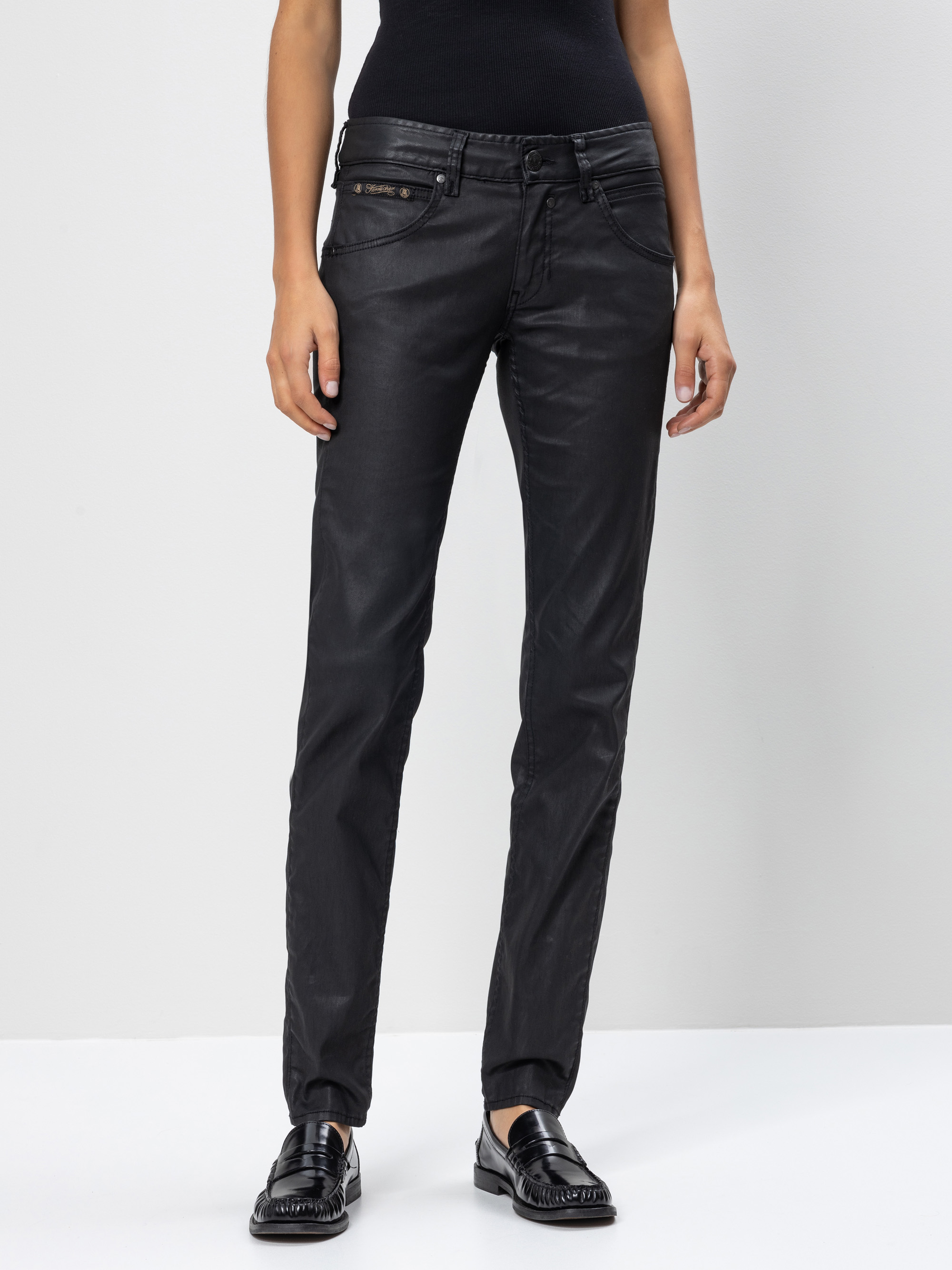 Eine Person, die ein schwarzes ärmelloses Oberteil, schwarze beschichtete Jeans und schwarze Halbschuhe trägt, steht vor einem hellgrauen Hintergrund. Das Foto ist von der Mitte des Oberkörpers bis zu den Füßen abgeschnitten.