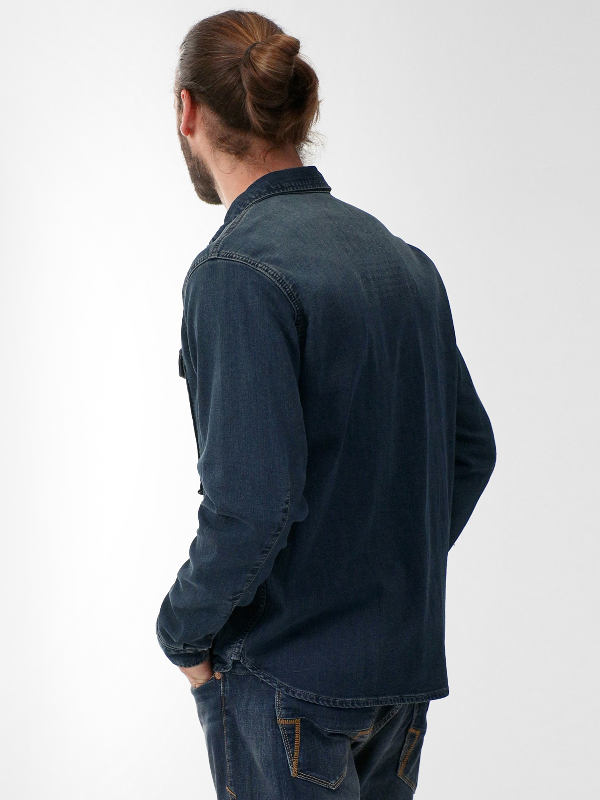 Ein Mann mit Bart und langem Haar in einem Dutt steht mit dem Rücken zur Kamera, trägt ein dunkles Jeanshemd und Jeans, eine Hand in der Tasche, vor einem schlichten hellen Hintergrund.