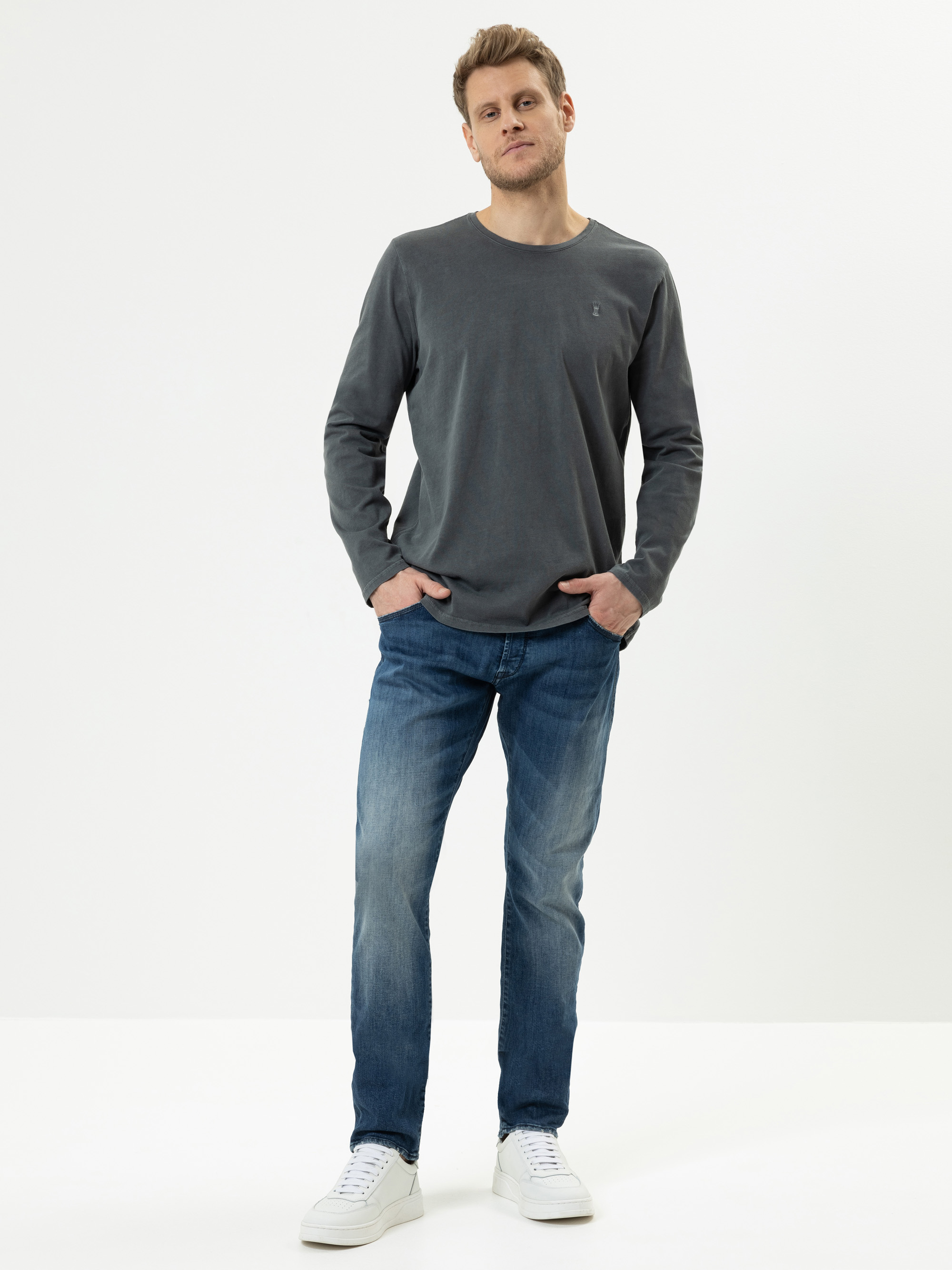 Ein Mann steht vor einem schlichten weißen Hintergrund und trägt ein langärmeliges graues Hemd, blaue Jeans und weiße Turnschuhe. Er hat die Hände in den Taschen und blickt leicht nach oben.