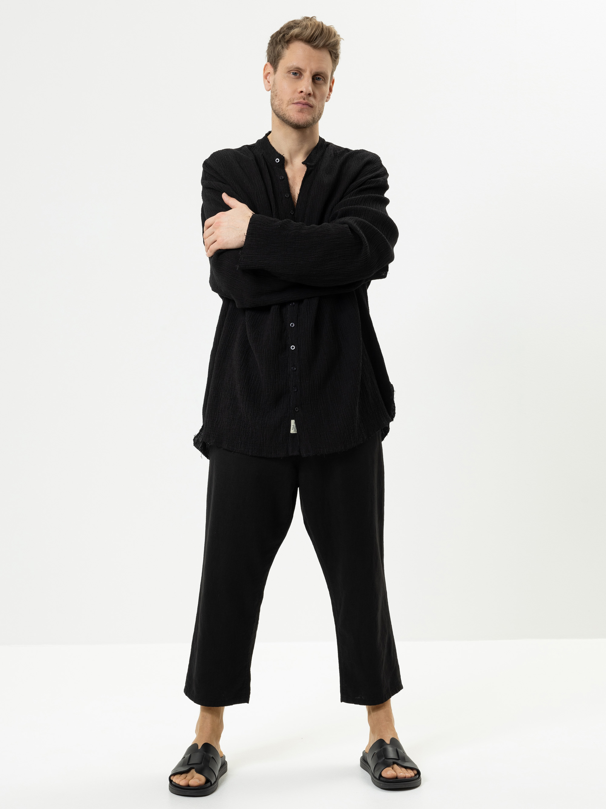 Ein Mann steht vor einem schlichten weißen Hintergrund, trägt ein lockeres schwarzes Button-up-Hemd, eine schwarze kurze Hose und schwarze Slide-Sandalen, hat die Arme verschränkt und einen neutralen Gesichtsausdruck.