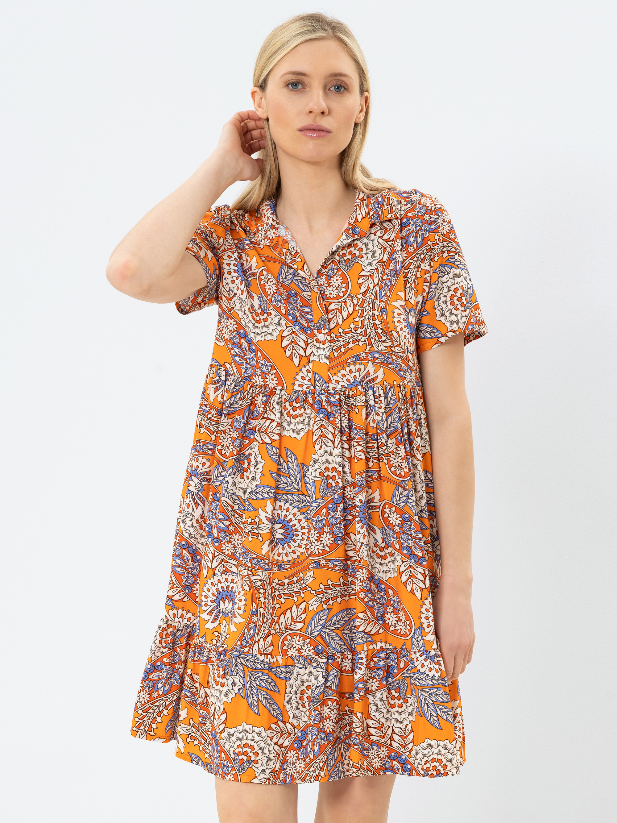 Eine Frau mit blondem Haar trägt ein orangefarbenes, kurzärmeliges, knielanges Kleid mit einem bunten Blumen- und Paisleymuster. Sie steht vor einem schlichten weißen Hintergrund und berührt mit einer Hand leicht ihr Haar.