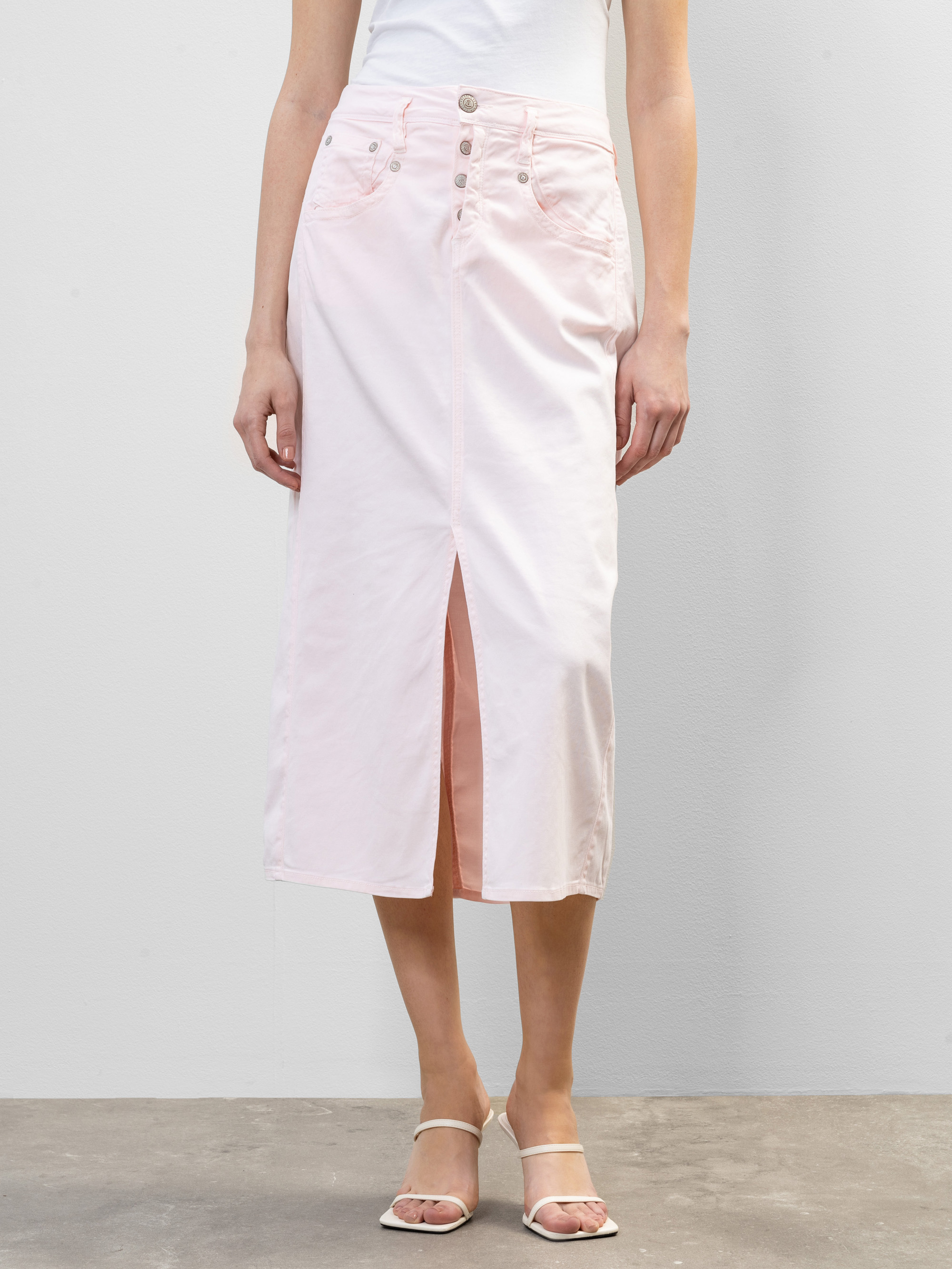 Eine Person, die ein weißes ärmelloses Oberteil, einen hellrosa Midirock mit hoher Taille, der vorne geknöpft und geschlitzt ist, und weiße hochhackige Sandalen trägt, steht vor einem hellgrauen Hintergrund.