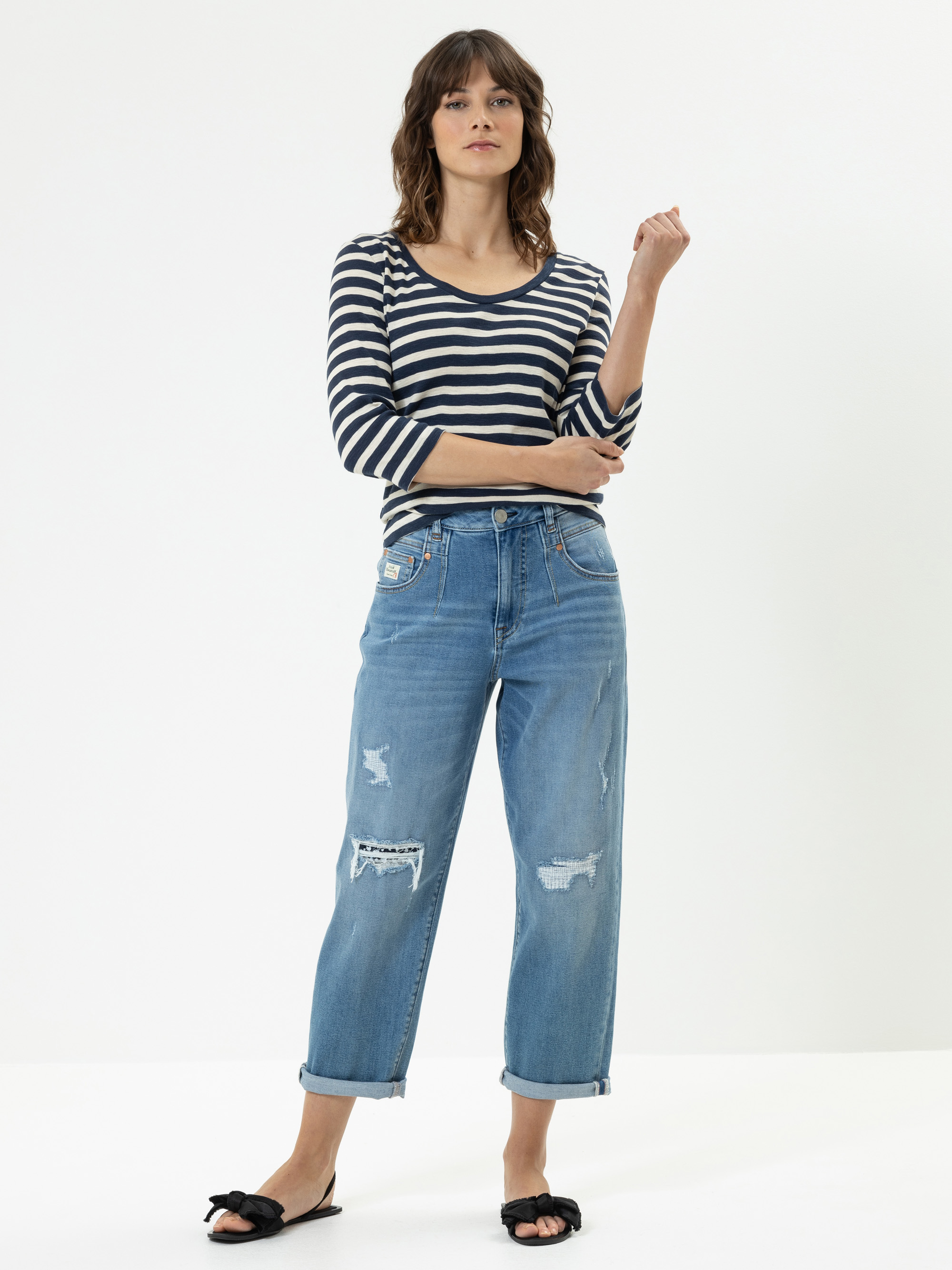 Eine Frau steht vor einem einfarbig weißen Hintergrund und trägt ein schwarz-weiß gestreiftes langärmeliges Oberteil, blaue zerrissene Jeans mit Bündchen und schwarze Slide-Sandalen mit Schleifen.