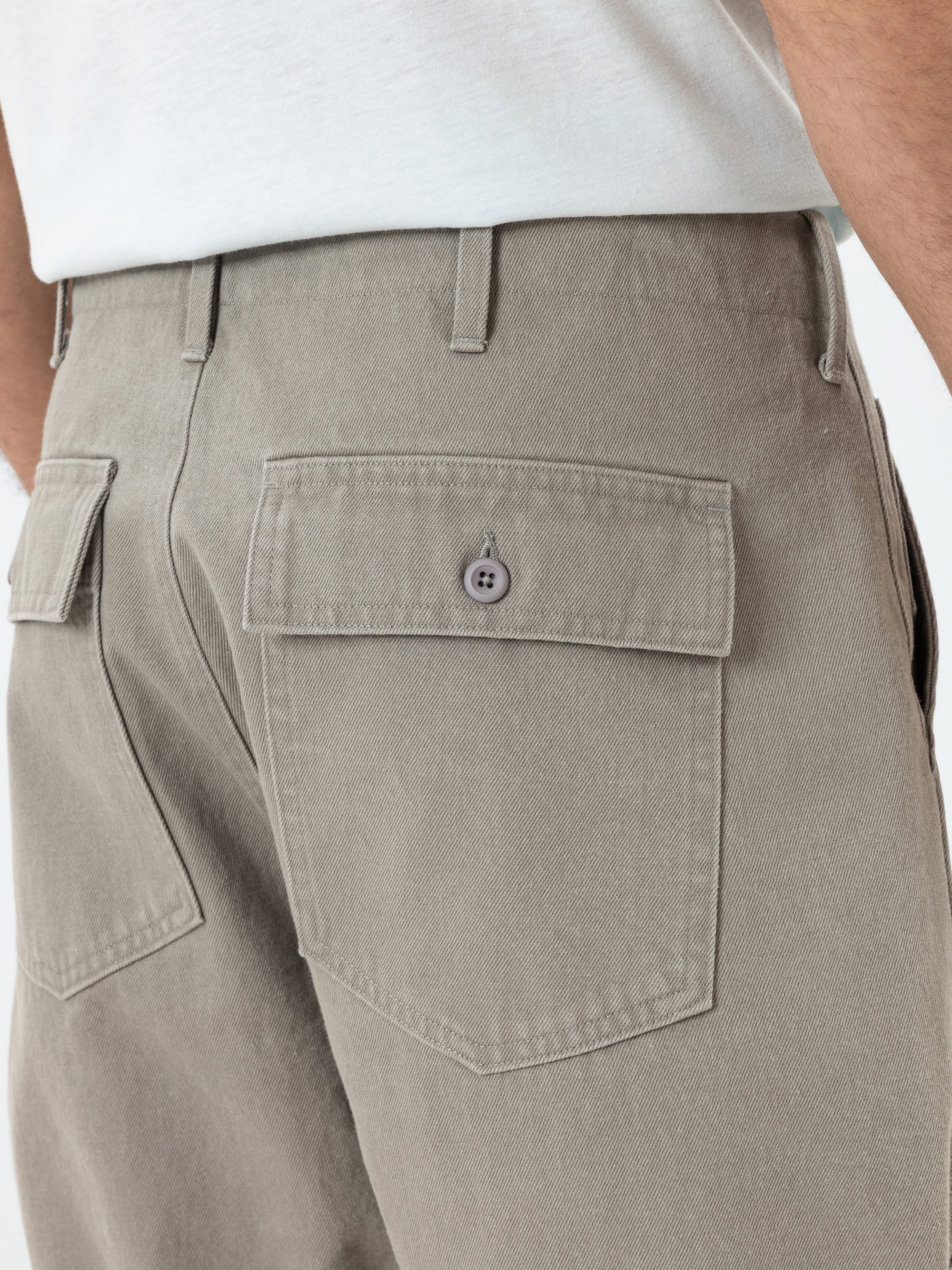 Nahaufnahme einer Person, die eine hellbraune Hose mit großen Gesäßtaschen trägt, von denen eine mit einem Knopf verschlossen ist, und ein weißes Hemd, das in die Hose gesteckt ist. Nur der untere Rücken und die Oberschenkel sind sichtbar.