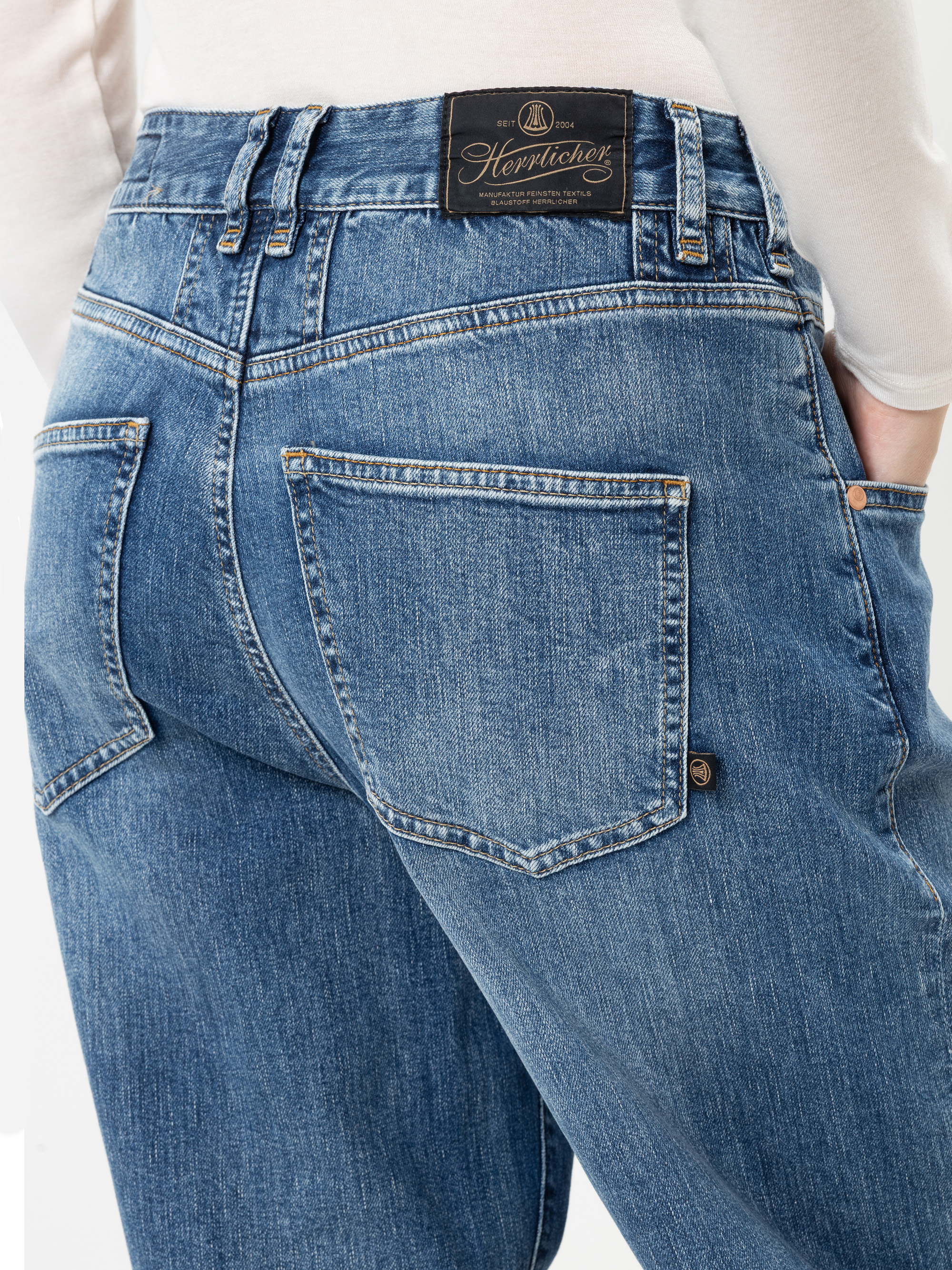 Eine Person, die blaue Jeans trägt, von hinten gesehen, mit sichtbaren Gesäßtaschen und einem schwarzen Leder-Markenetikett am Hosenbund. Die Person trägt außerdem ein weißes Oberteil.