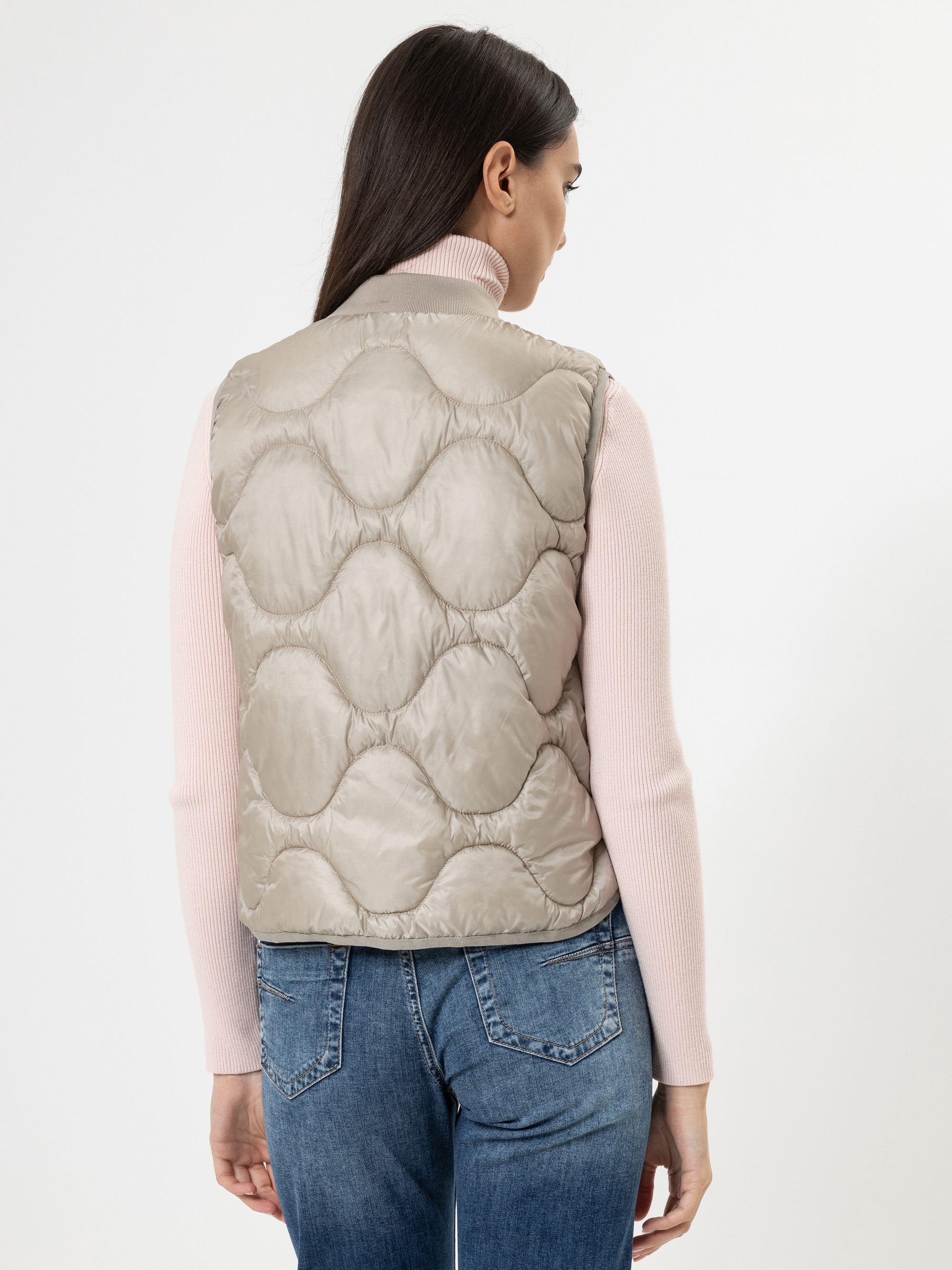 Man sieht eine Frau mit langen braunen Haaren von hinten, die eine hellbeige Steppweste über einem blassrosa Rollkragenpullover und blaue Jeans trägt und vor einem schlichten weißen Hintergrund steht.