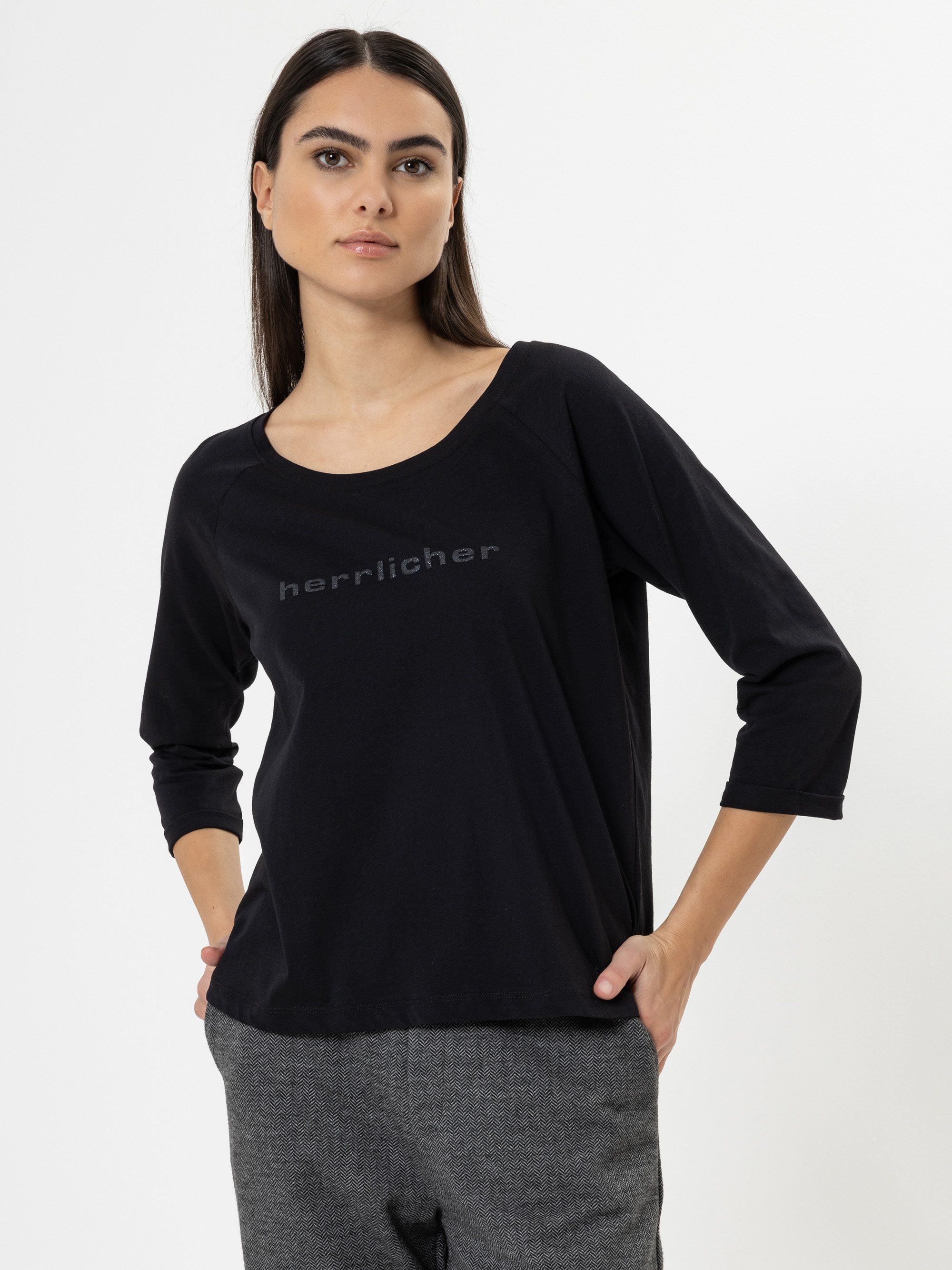 Eine Frau mit langen dunklen Haaren trägt ein schwarzes langärmeliges Hemd mit herrlicher Aufschrift auf der Vorderseite und eine graue Hose und steht mit einer Hand in der Tasche vor einem schlichten weißen Hintergrund.