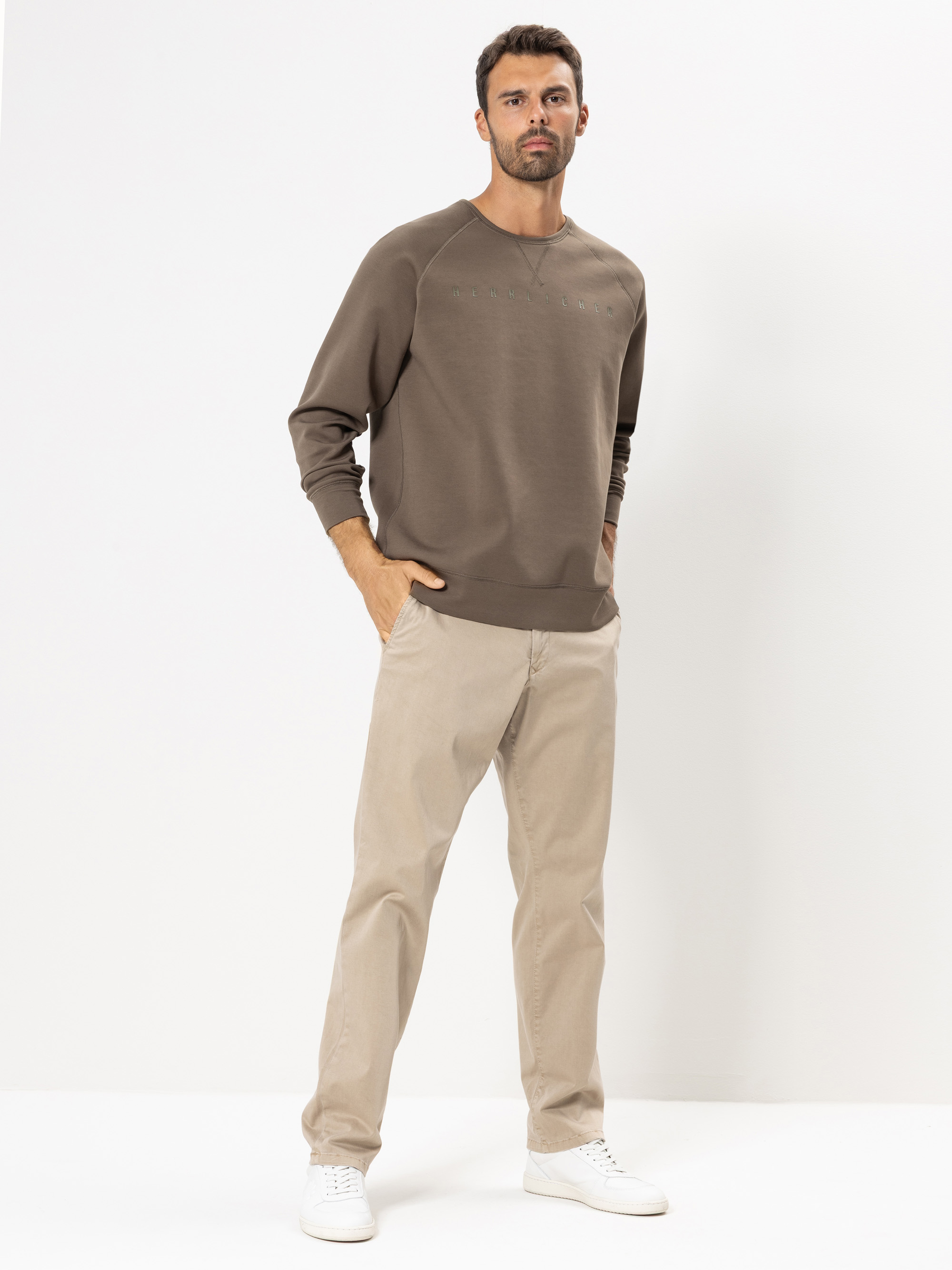 Ein Mann steht vor einem schlichten weißen Hintergrund und trägt ein braunes langärmeliges Sweatshirt, eine beigefarbene Hose und weiße Turnschuhe. Seine Hände stecken in den Hosentaschen, und er blickt selbstbewusst in die Kamera.