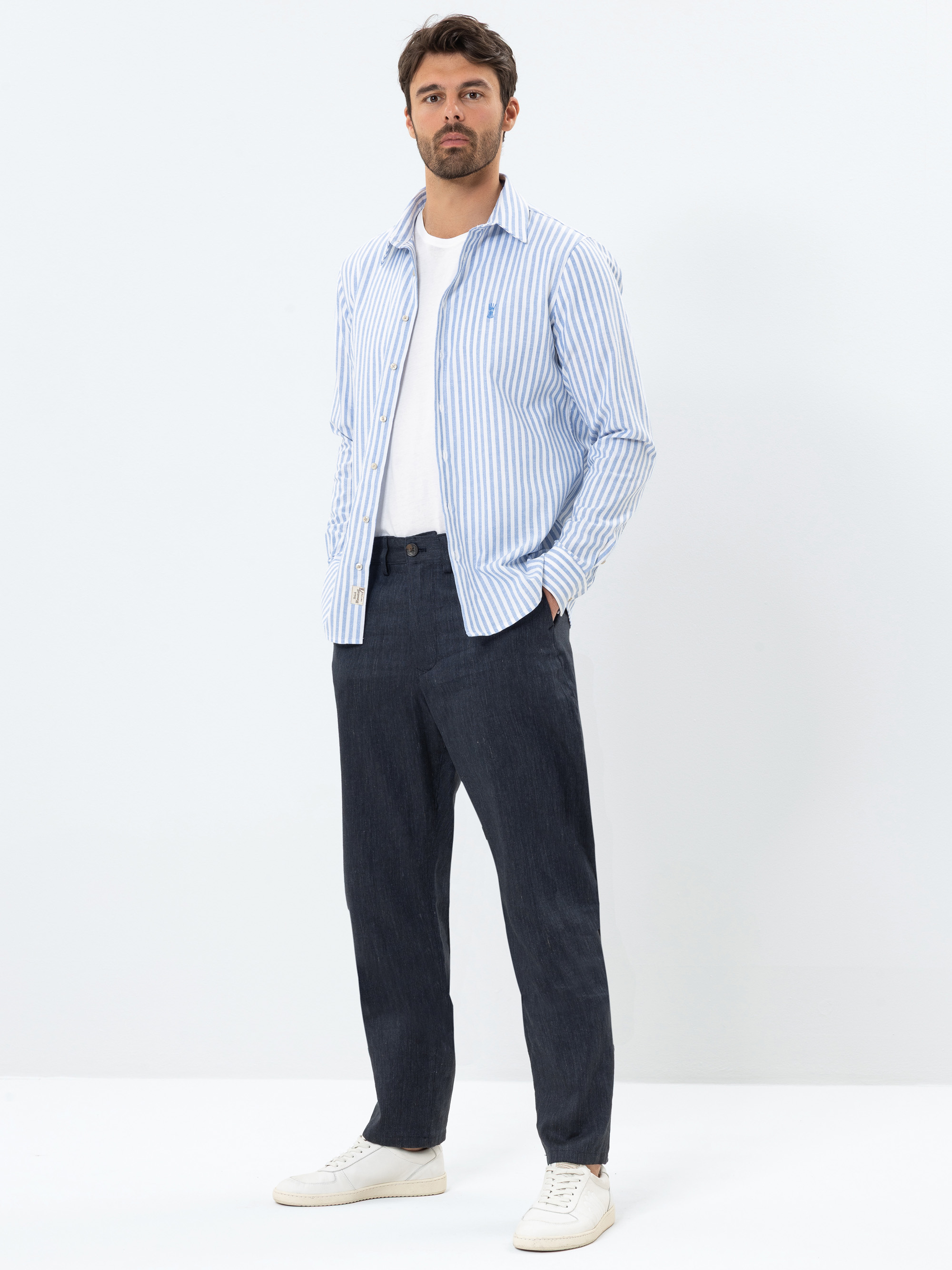 Ein Mann steht vor einem schlichten weißen Hintergrund und trägt ein blau-weiß gestreiftes Button-down-Hemd über einem weißen T-Shirt, eine dunkle Hose und weiße Turnschuhe. Er hat kurzes dunkles Haar und einen Bart und hat die Hände in den Taschen