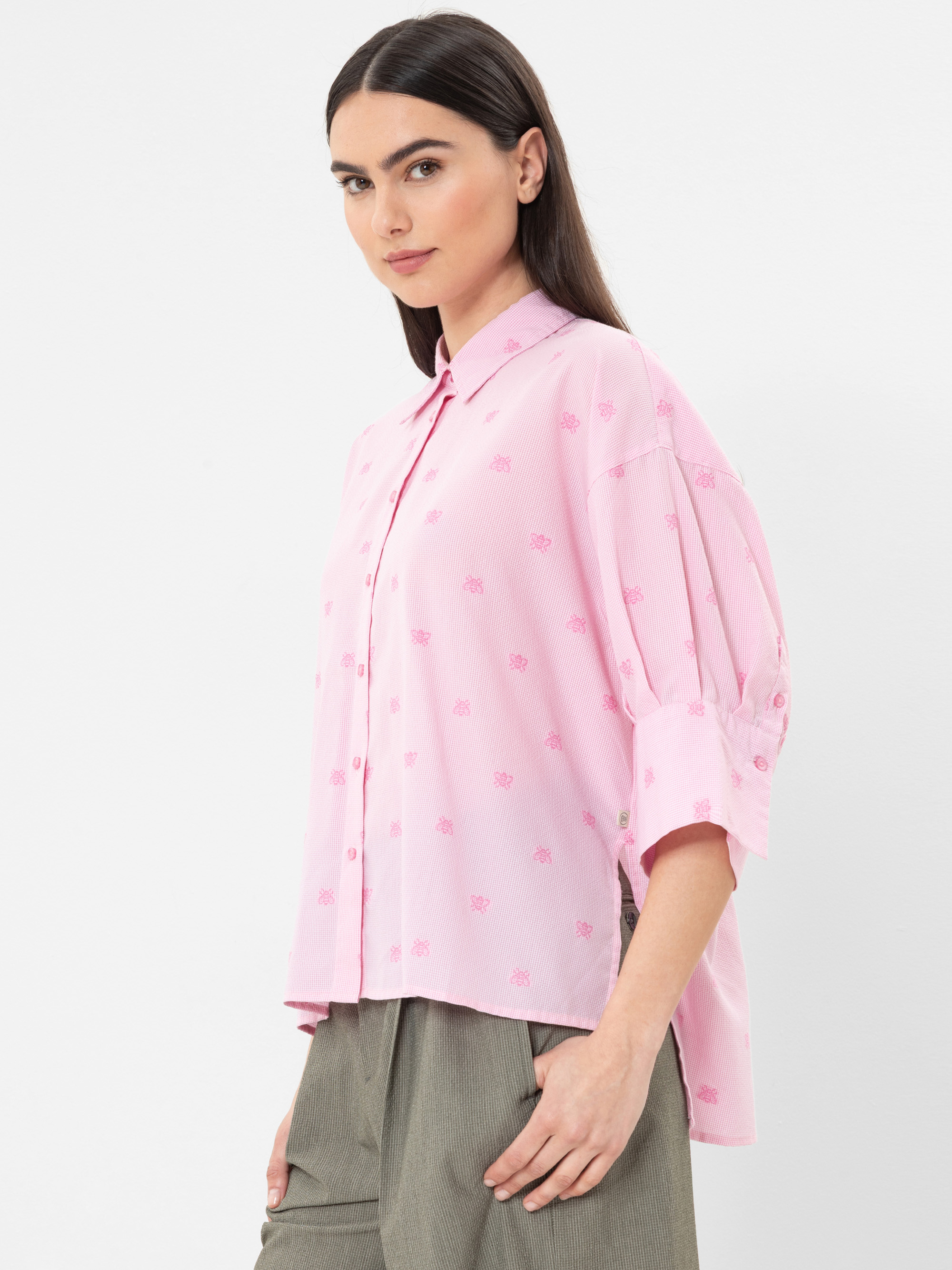 Eine Frau in einem hellrosafarbenen, locker sitzenden Button-up-Hemd mit dezentem Blumenmuster und ellbogenlangen Ärmeln, gepaart mit einer olivgrünen Hose, steht vor einem schlichten weißen Hintergrund.