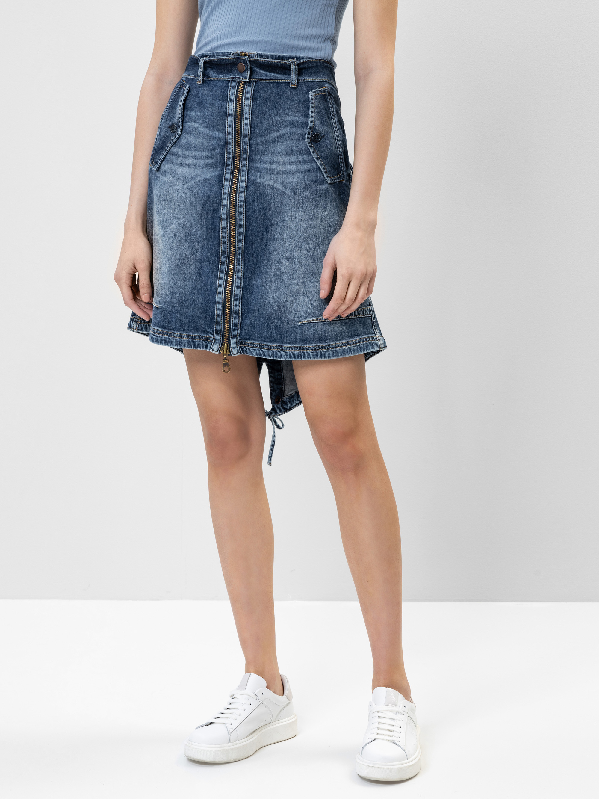 Eine Person, die ein blaues geripptes ärmelloses Oberteil, einen Jeansrock mit Taschen und weiße Turnschuhe trägt, steht vor einem schlichten hellgrauen Hintergrund.
