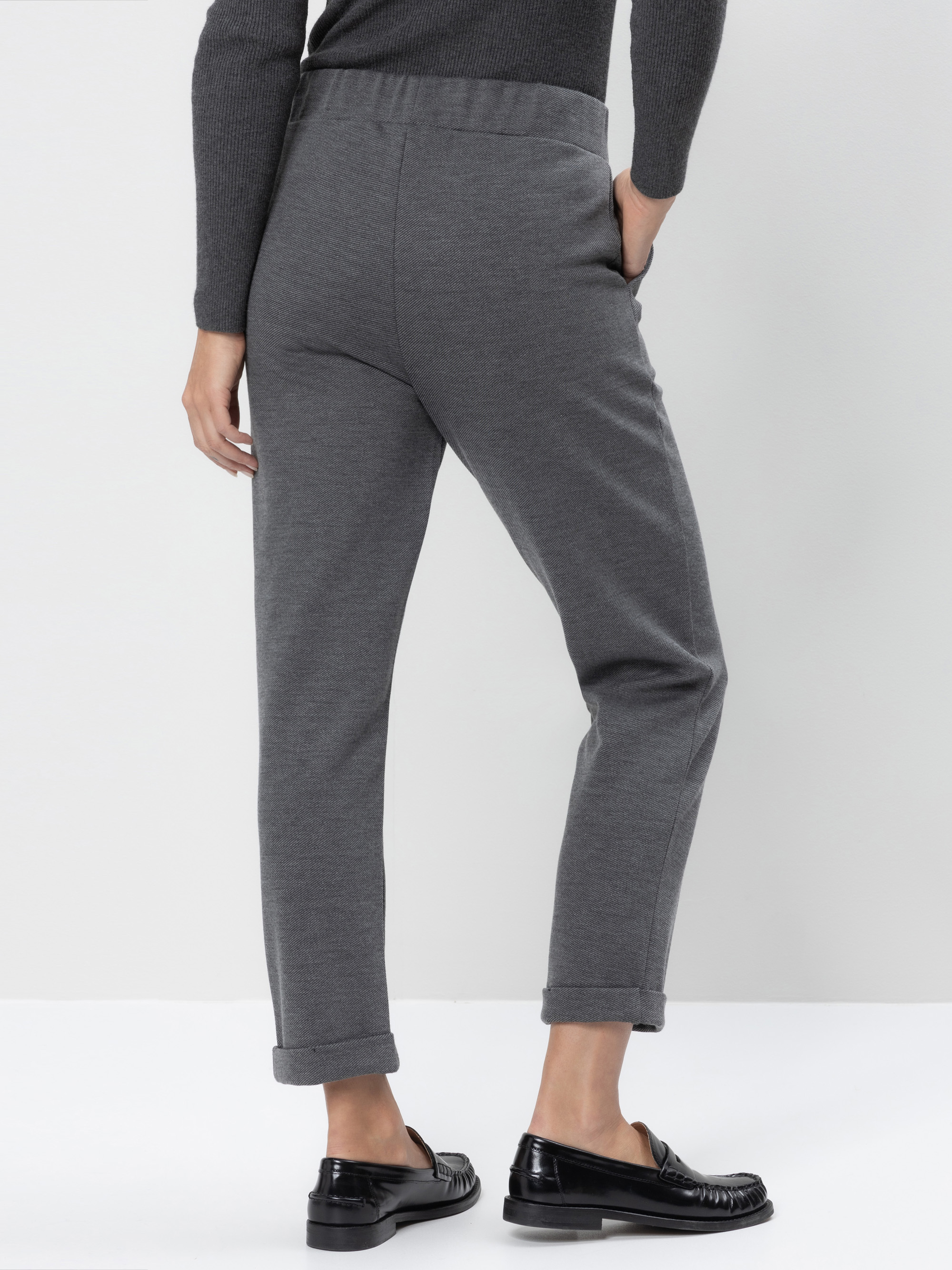 Person, die vor einem weißen Hintergrund steht und eine graue Hose mit Manschetten, ein passendes graues langärmeliges Oberteil und schwarze Halbschuhe trägt. Die linke Hand steckt in der Hosentasche, der Blick ist von der Kamera abgewandt.