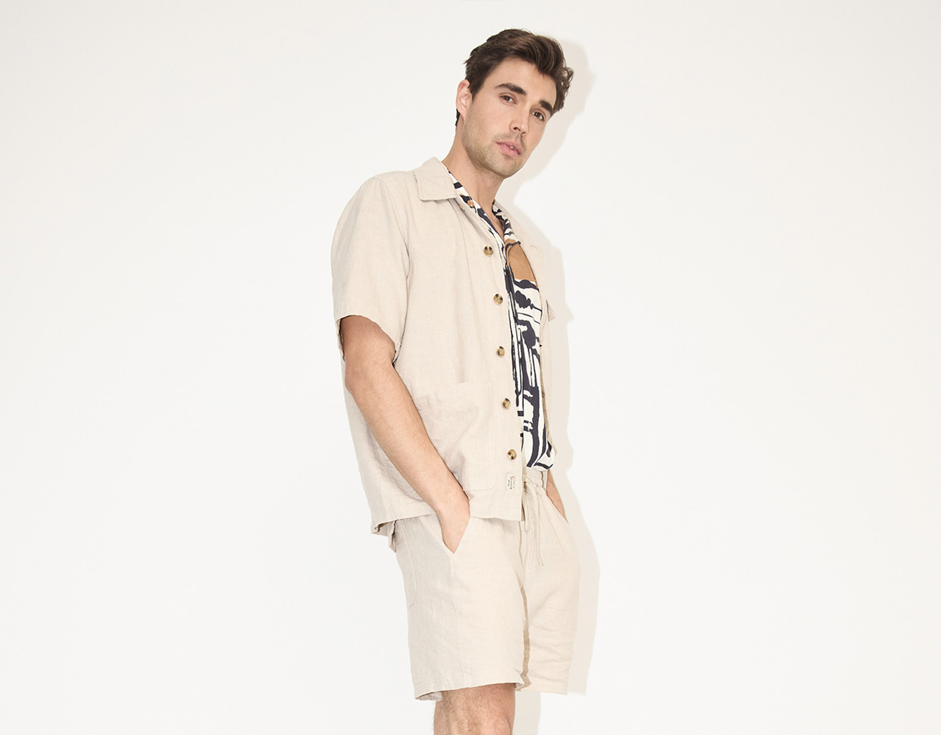 Ein Mann steht vor einem schlichten weißen Hintergrund und trägt ein hellbeiges kurzärmeliges Hemd, passende Shorts und darunter ein gemustertes Hemd. Er hat die Hände in den Taschen und blickt selbstbewusst in die Kamera.