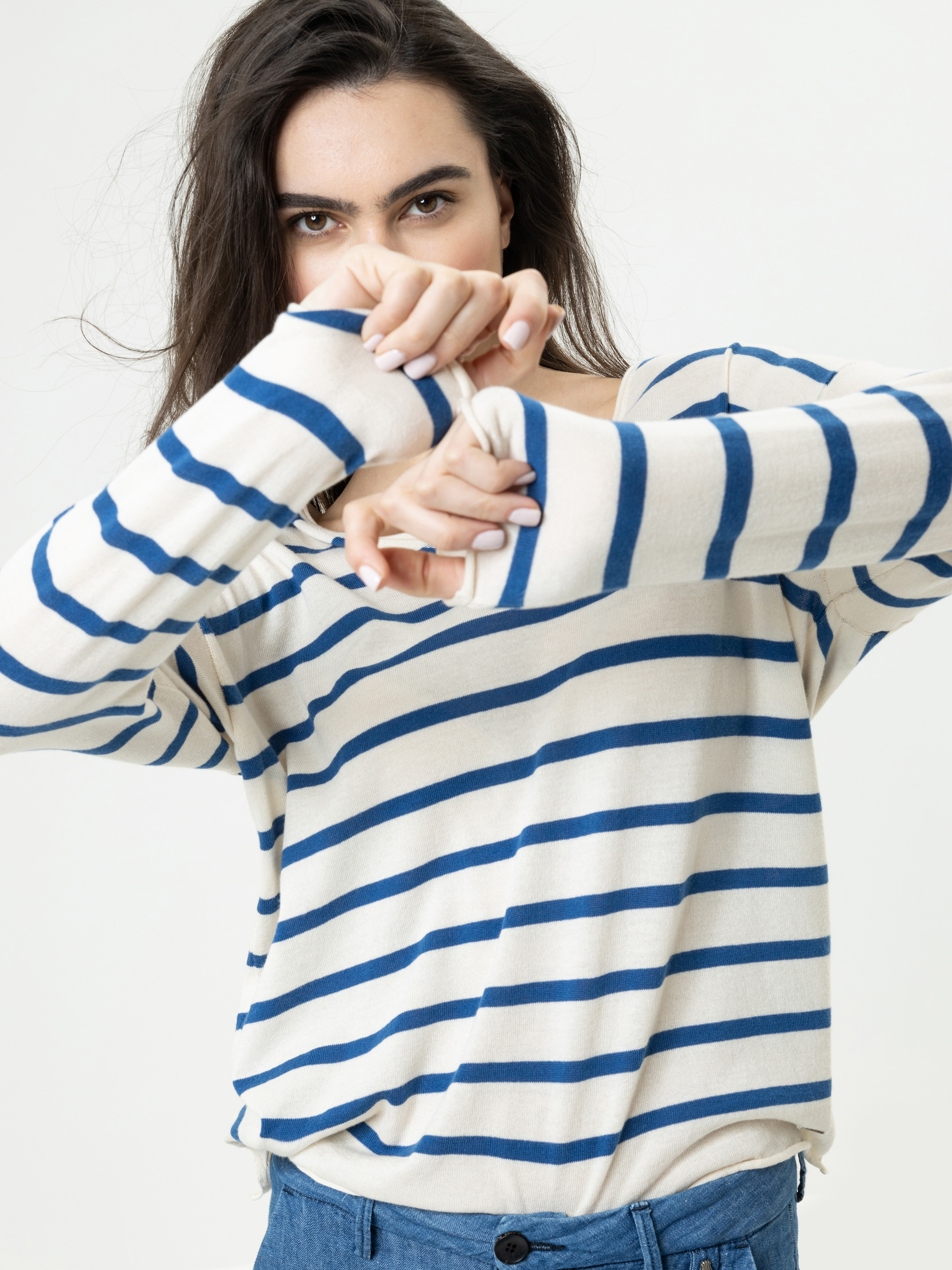 Eine Person, die ein cremefarbenes und blau gestreiftes langärmeliges Hemd trägt, posiert mit vor dem Gesicht verschränkten Armen und blickt direkt in die Kamera, wobei der Hintergrund neutral ist.