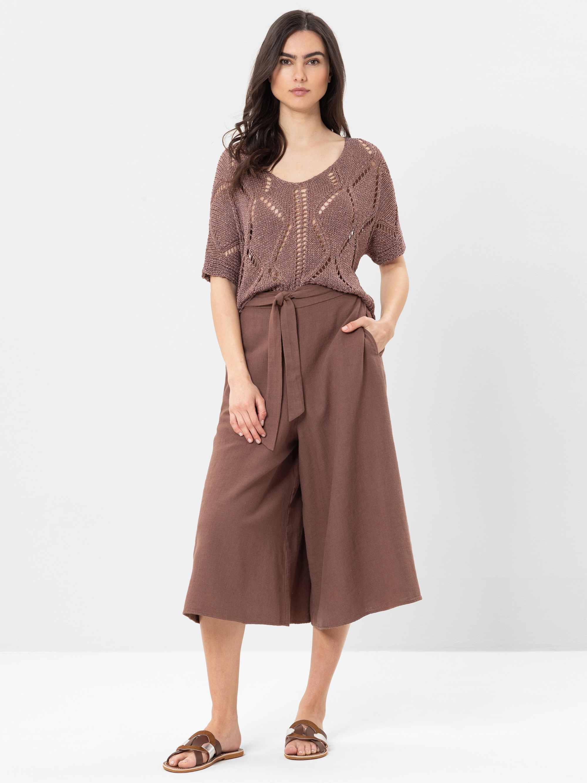 Eine Frau steht vor einem unifarbenen Hintergrund und trägt ein mauvefarbenes, kurzärmeliges Strickoberteil, eine braune, weit geschnittene Culotte-Hose mit Bindeband und braune Gleitsandalen. Sie hat langes dunkles Haar und einen neutralen Gesichtsausd