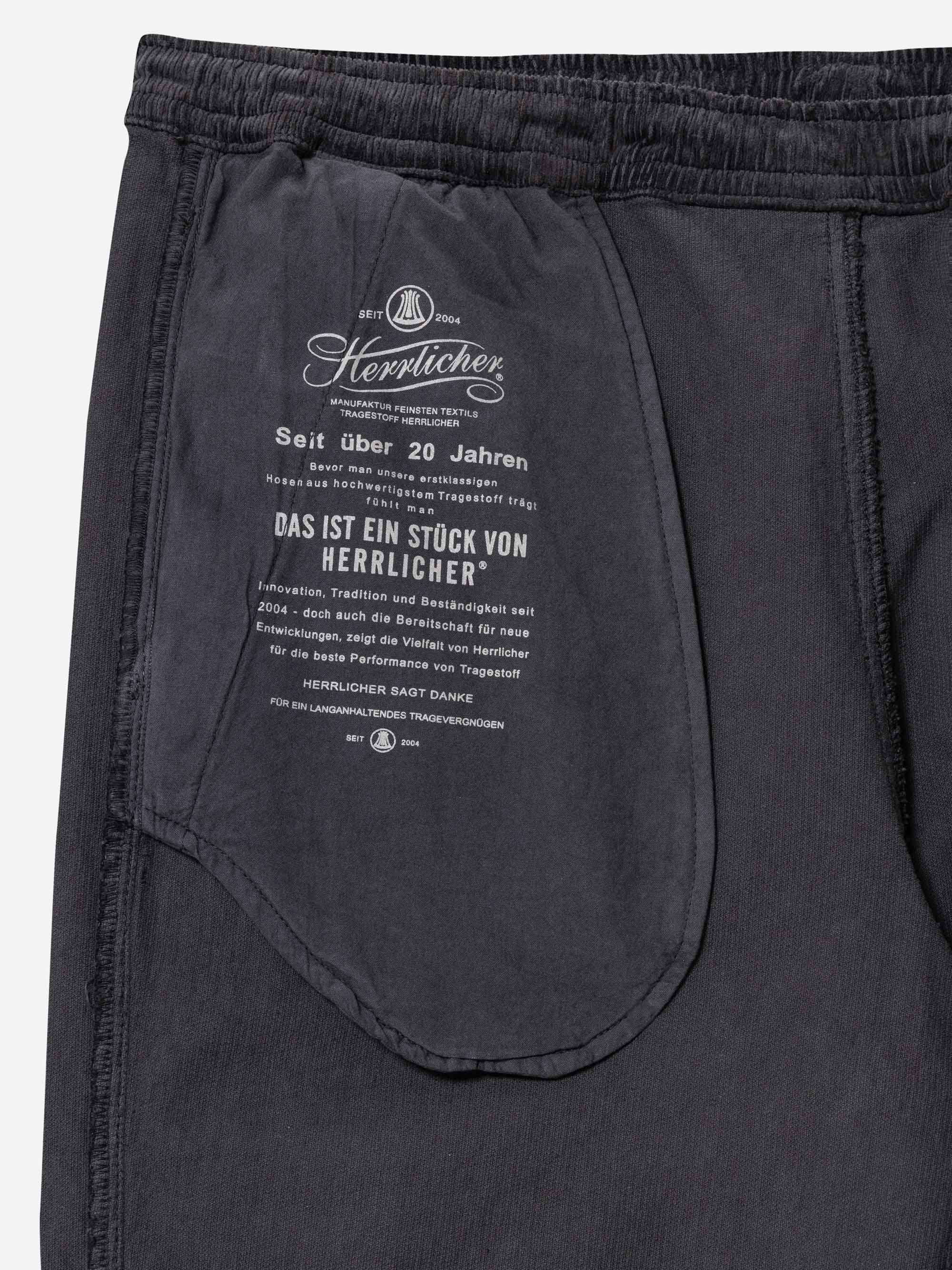 Nahaufnahme eines schwarzen Kleidungsstücks mit einer Innentasche, auf der ein weißer Text und ein Logo in deutscher Sprache aufgedruckt sind, das auf Herlicher hinweist und Informationen über die Tradition und Qualität der Marke enthält.