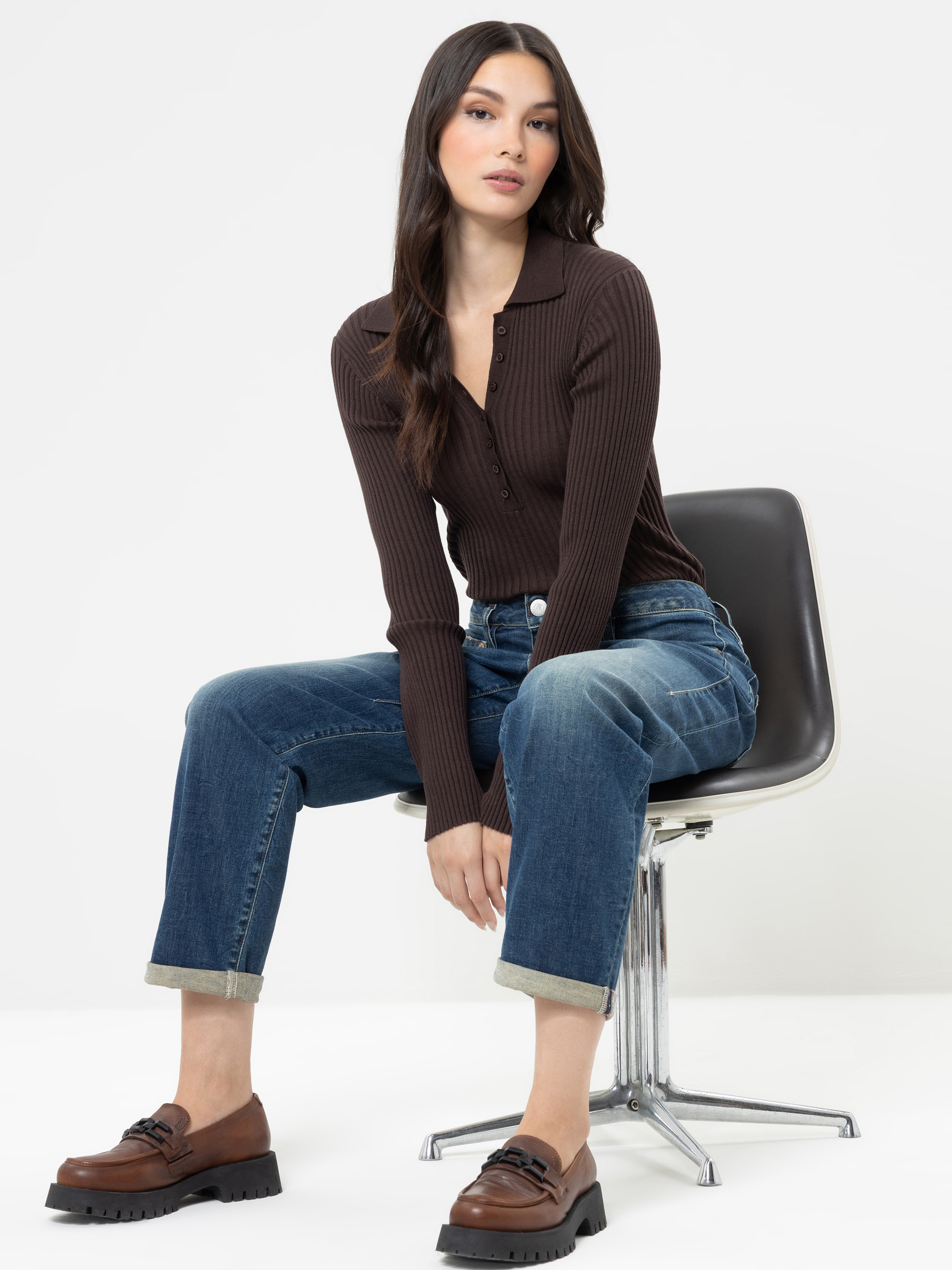 Eine Frau mit langen dunklen Haaren sitzt auf einem modernen Stuhl und trägt ein dunkelbraunes, langärmeliges, geripptes Oberteil, eine blaue Jeans mit Umschlag und braune Slipper. Vor einem schlichten weißen Hintergrund blickt sie mit einer entspannte