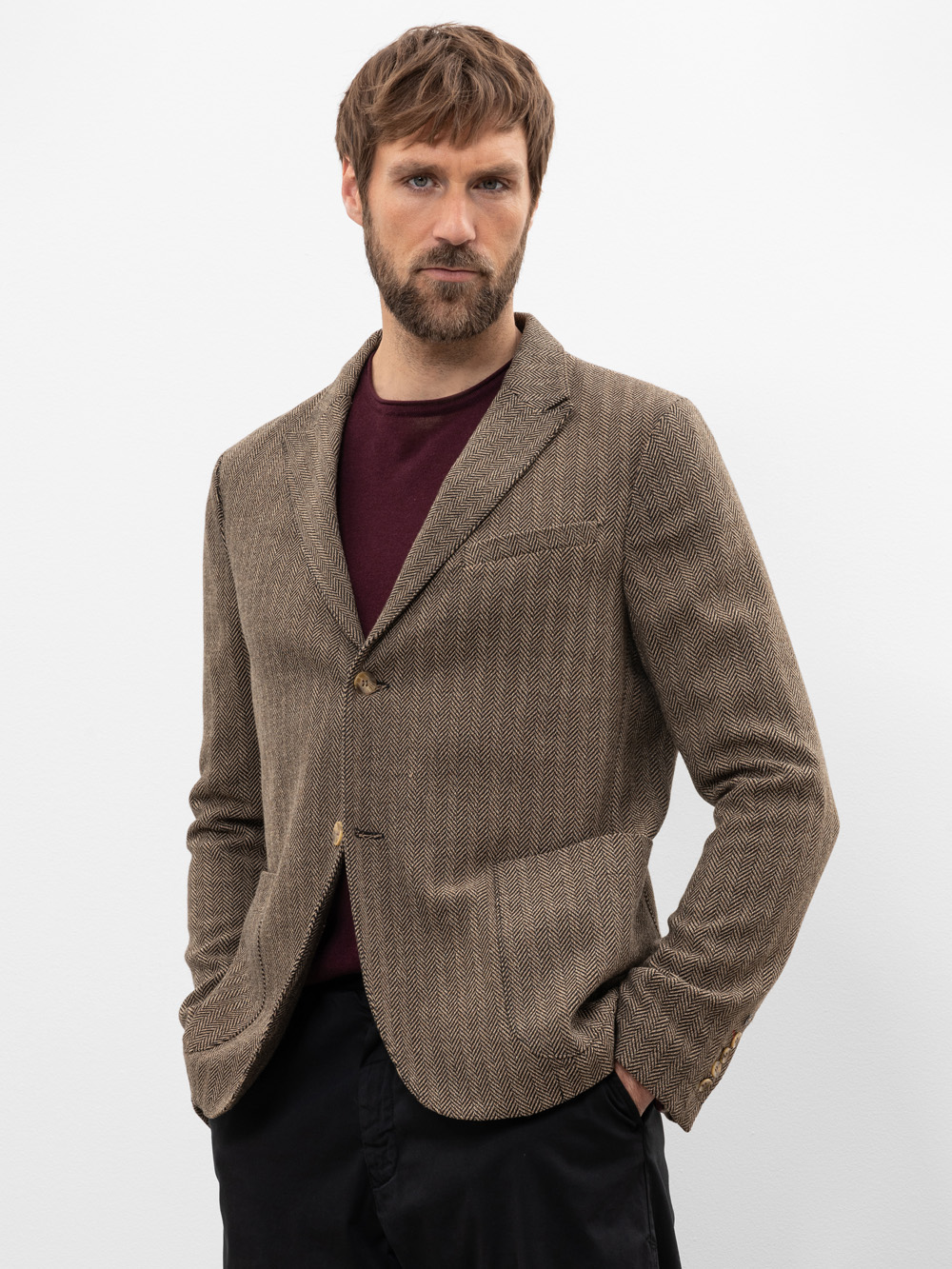 Ein Mann mit kurzen braunen Haaren und einem Bart trägt einen braunen strukturierten Blazer über einem dunkelkastanienbraunen Hemd und einer schwarzen Hose. Er steht vor einem schlichten weißen Hintergrund und hat die Hände in den Taschen.