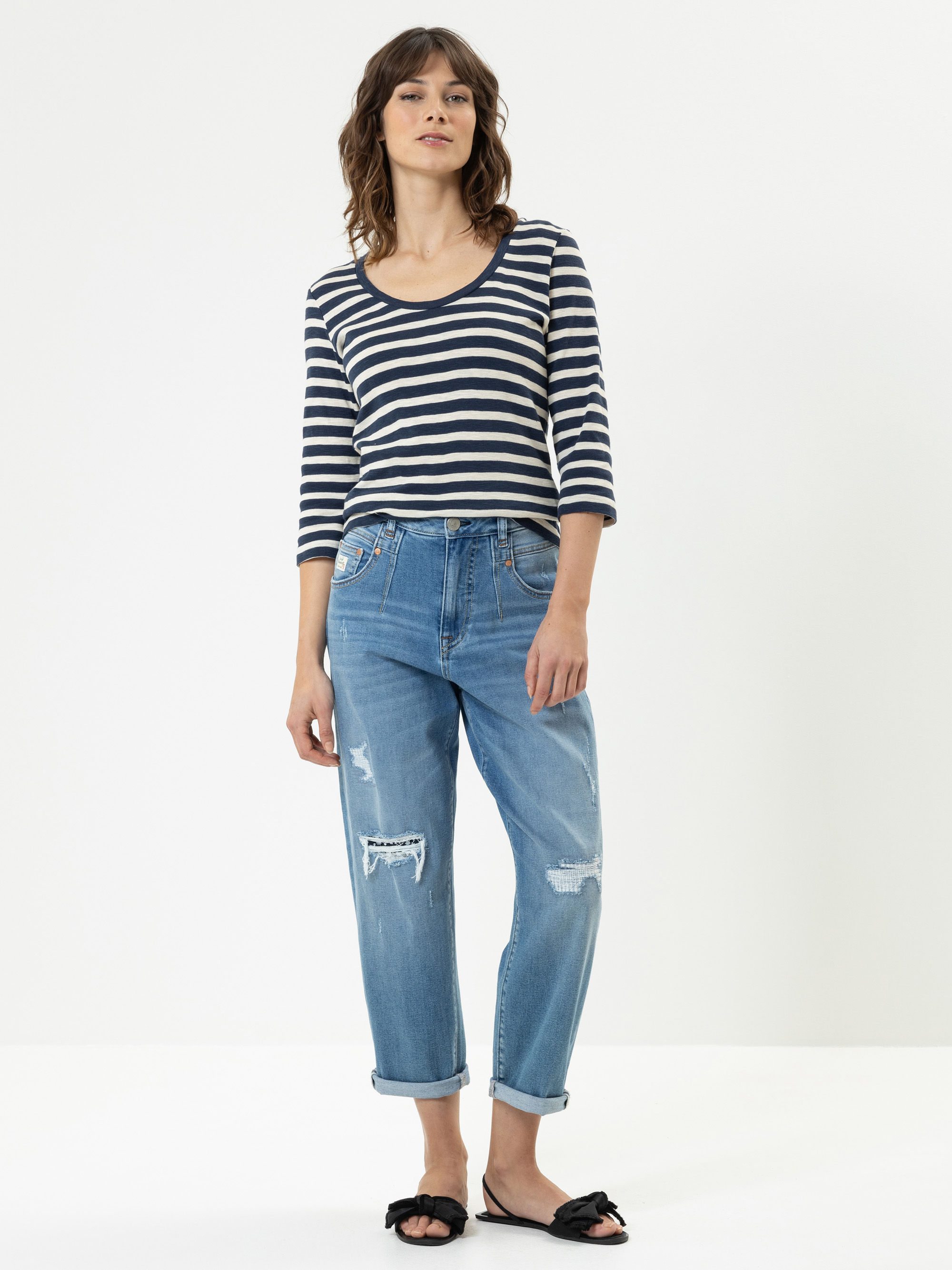 Eine Frau steht vor einem weißen Hintergrund und trägt ein marineblau-weiß gestreiftes, langärmeliges Oberteil, abgenutzte Bluejeans mit aufgerollten Manschetten und schwarze Slipper-Sandalen. Sie hat gewelltes braunes Haar und einen entspannten Gesic