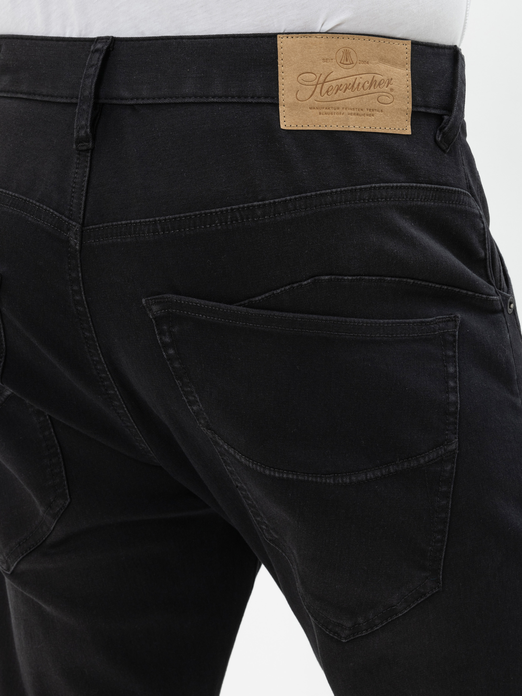 Nahaufnahme der Rückseite einer schwarzen Jeans mit zwei großen Gesäßtaschen und einem braunen Lederaufnäher am Bund mit der Aufschrift Herrlicher in Schreibschrift. Die Jeans wird mit einem weißen Hemd in der Taille getragen.