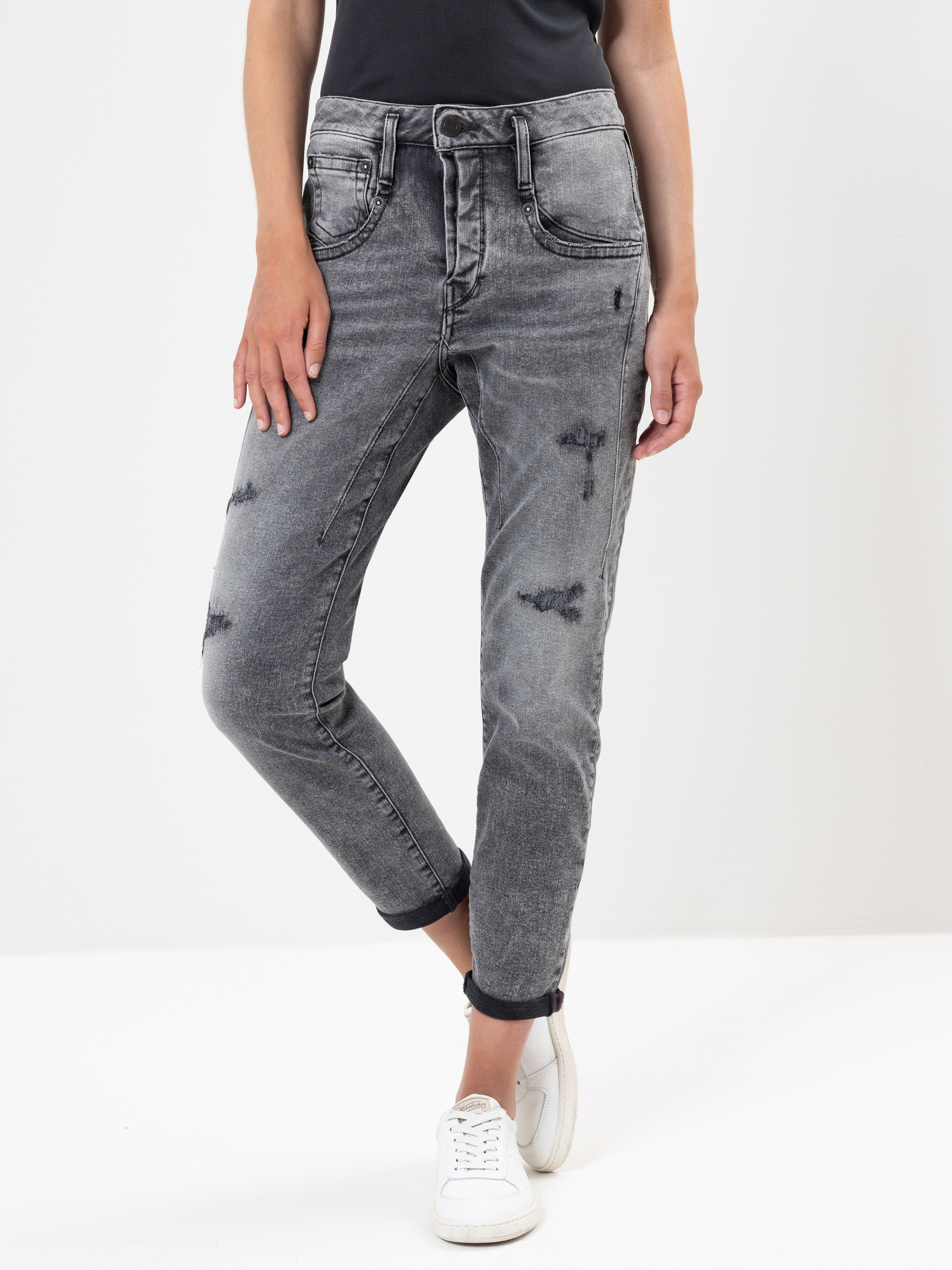 Eine Person, die ein schwarzes Oberteil, verwaschene graue Jeans mit aufgerollten Bündchen und weiße Turnschuhe trägt, steht vor einem schlichten weißen Hintergrund. Ihre Hände ruhen an den Seiten.