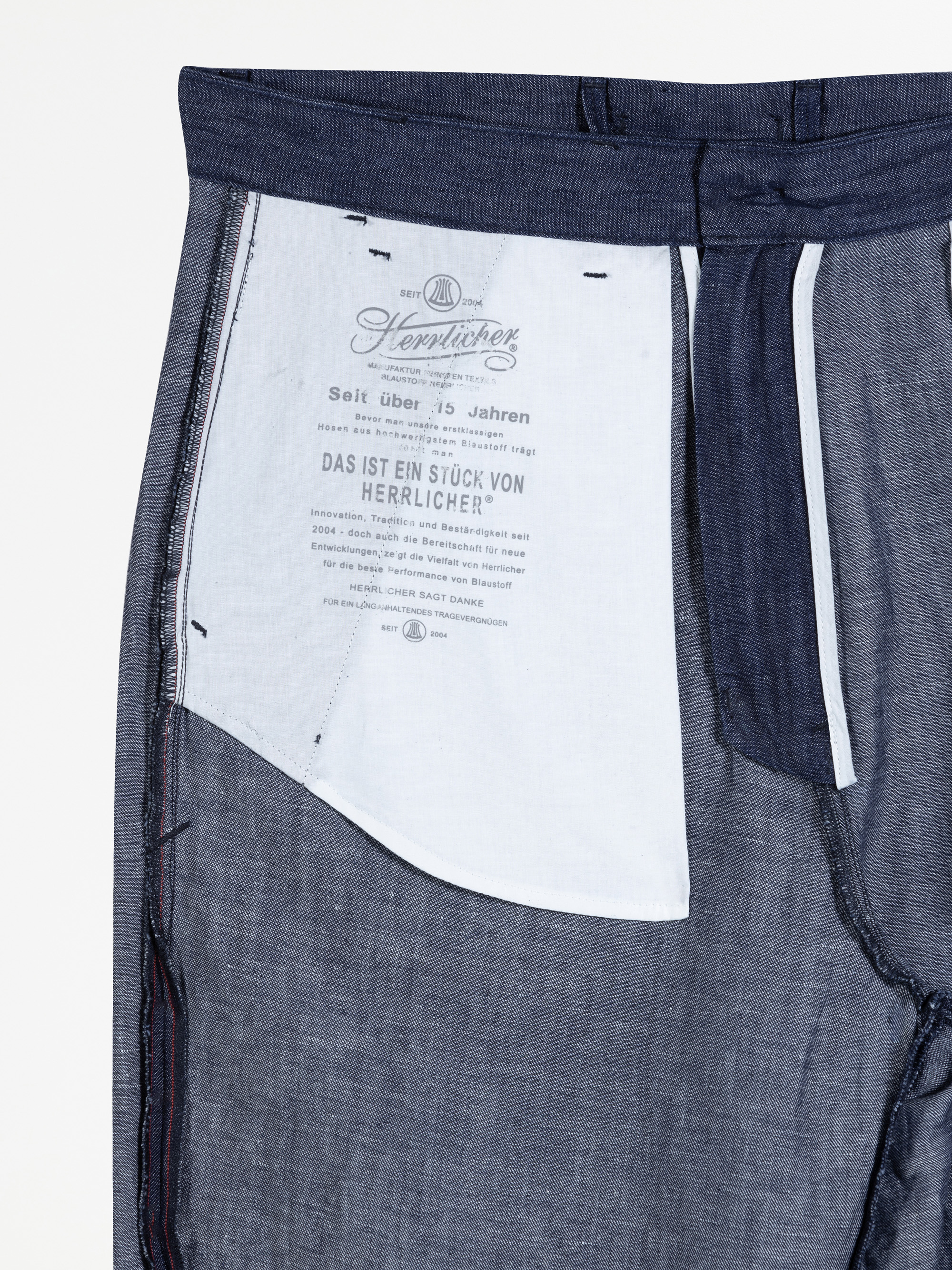 Nahaufnahme der Innenseite einer blauen Jeans, die das weiße Innenfutter der Taschen mit aufgedrucktem Text in deutscher Sprache und dem Markennamen Herrlicher zeigt.