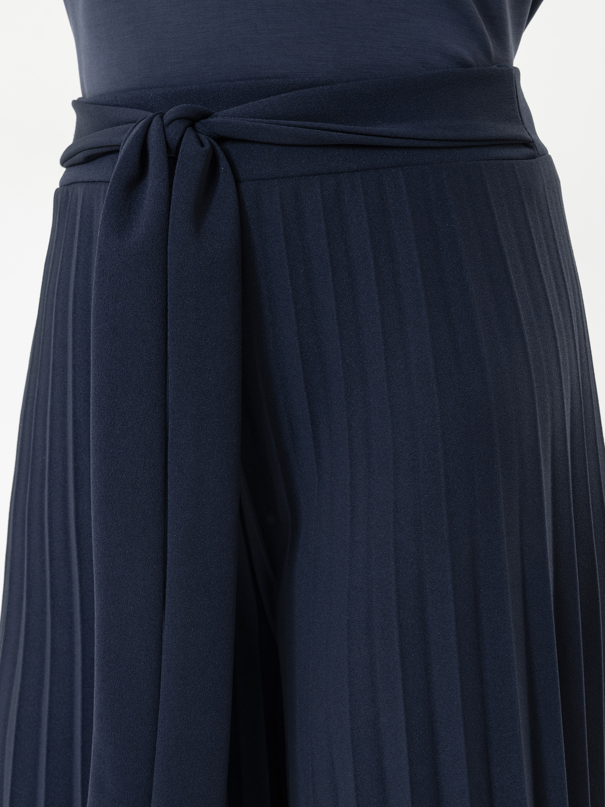 Nahaufnahme eines dunkelblauen Faltenrocks oder einer Hose mit einem breiten, passenden Stoffgürtel, der in der Taille mit einem einfachen Knoten gebunden wird. Der Stoff hat eine weiche Textur und vertikale Falten.