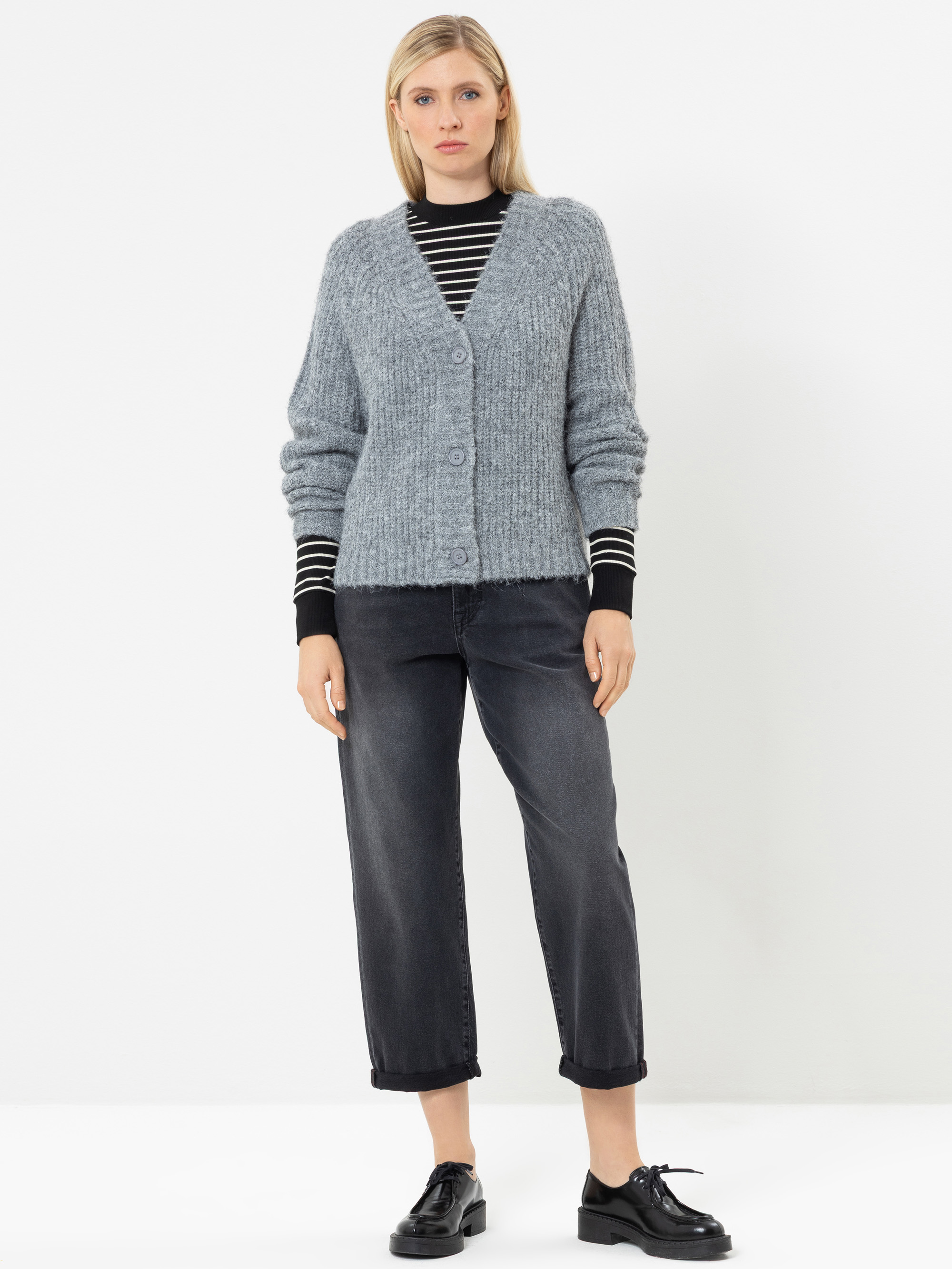 Eine Frau steht vor einem weißen Hintergrund und trägt eine graue Strickjacke mit Knöpfen über einem schwarz-weiß gestreiften Oberteil, eine dunkle, kurz geschnittene Jeans und schwarze Schuhe.