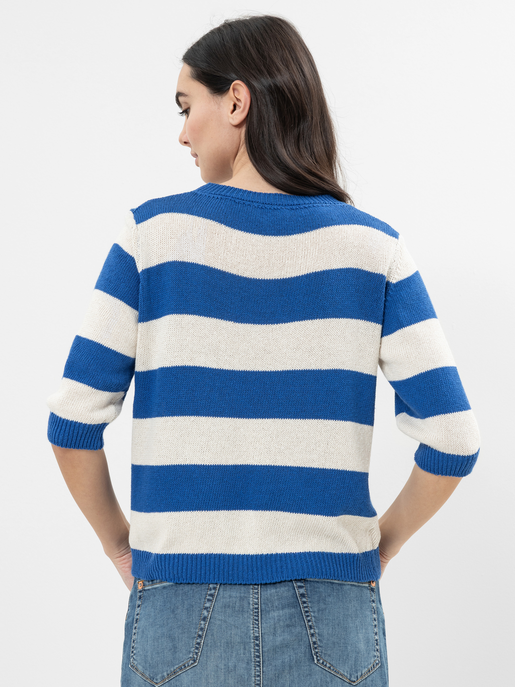 Eine Frau mit langen dunklen Haaren wird von hinten gezeigt. Sie trägt einen blau-weiß gestreiften Pullover mit ellenbogenlangen Ärmeln und blaue Jeans. Sie steht vor einem schlichten weißen Hintergrund.