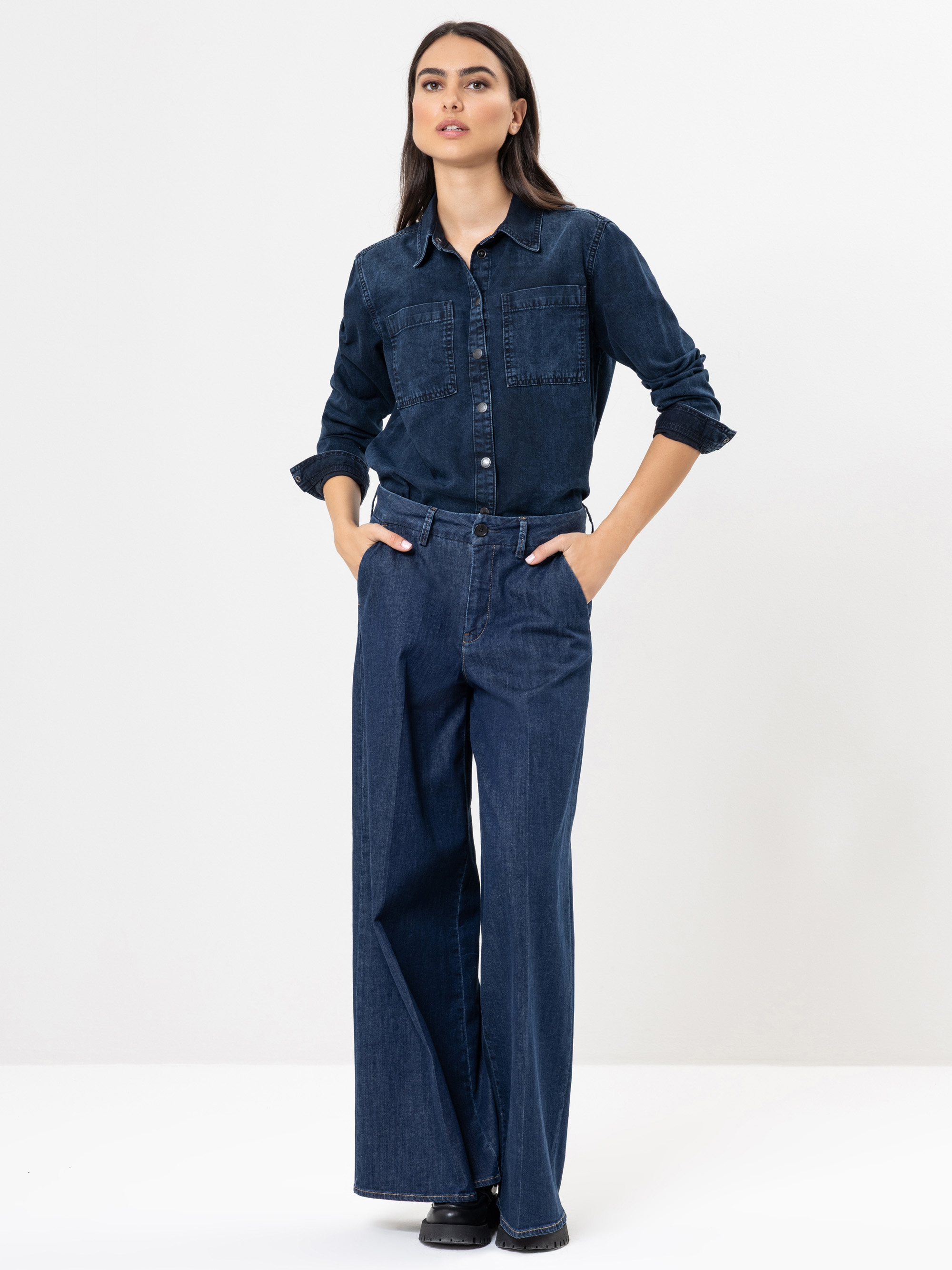 Eine Frau steht vor einem schlichten weißen Hintergrund und trägt ein dunkles Jeanshemd mit gekrempelten Ärmeln und eine weit geschnittene Jeans. Sie hat die Hände in den Taschen und trägt schwarze Schuhe.