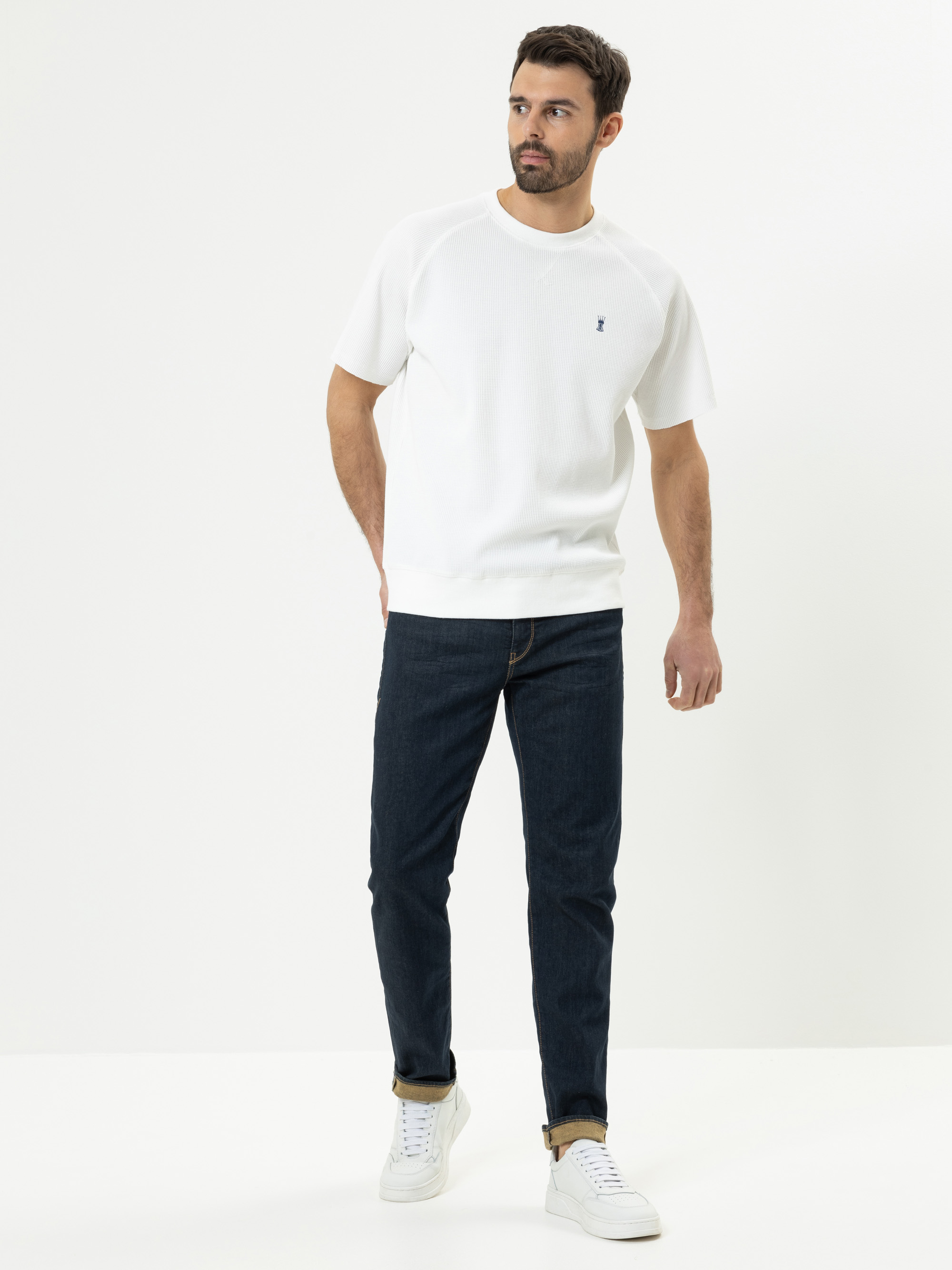 Ein Mann steht vor einem schlichten weißen Hintergrund und trägt ein weißes kurzärmeliges T-Shirt, dunkelblaue Jeans und weiße Turnschuhe. Er blickt leicht nach links und nimmt eine entspannte, lässige Haltung ein.