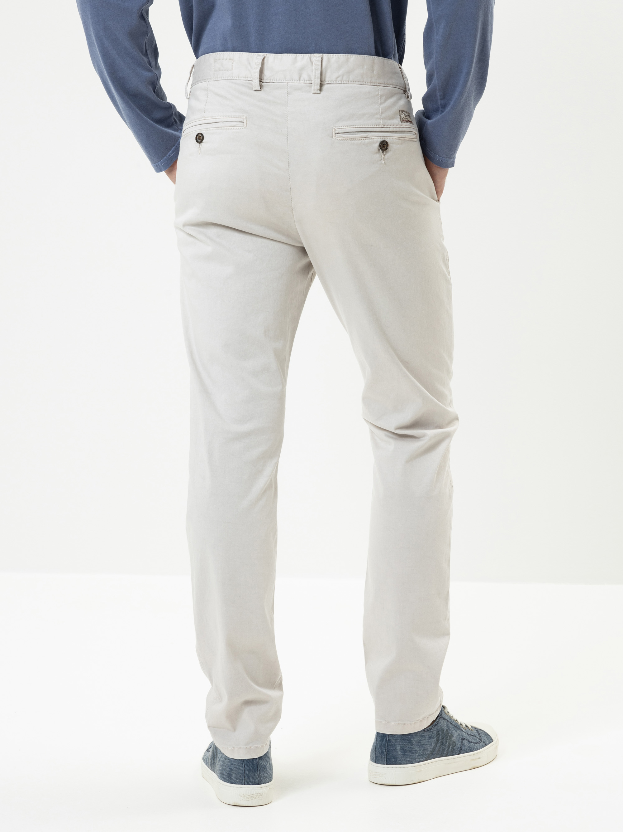 Es wird eine Person von hinten gezeigt, die eine hellbeige Hose, ein blaues langärmeliges Hemd und blaue Turnschuhe vor einem schlichten weißen Hintergrund trägt.