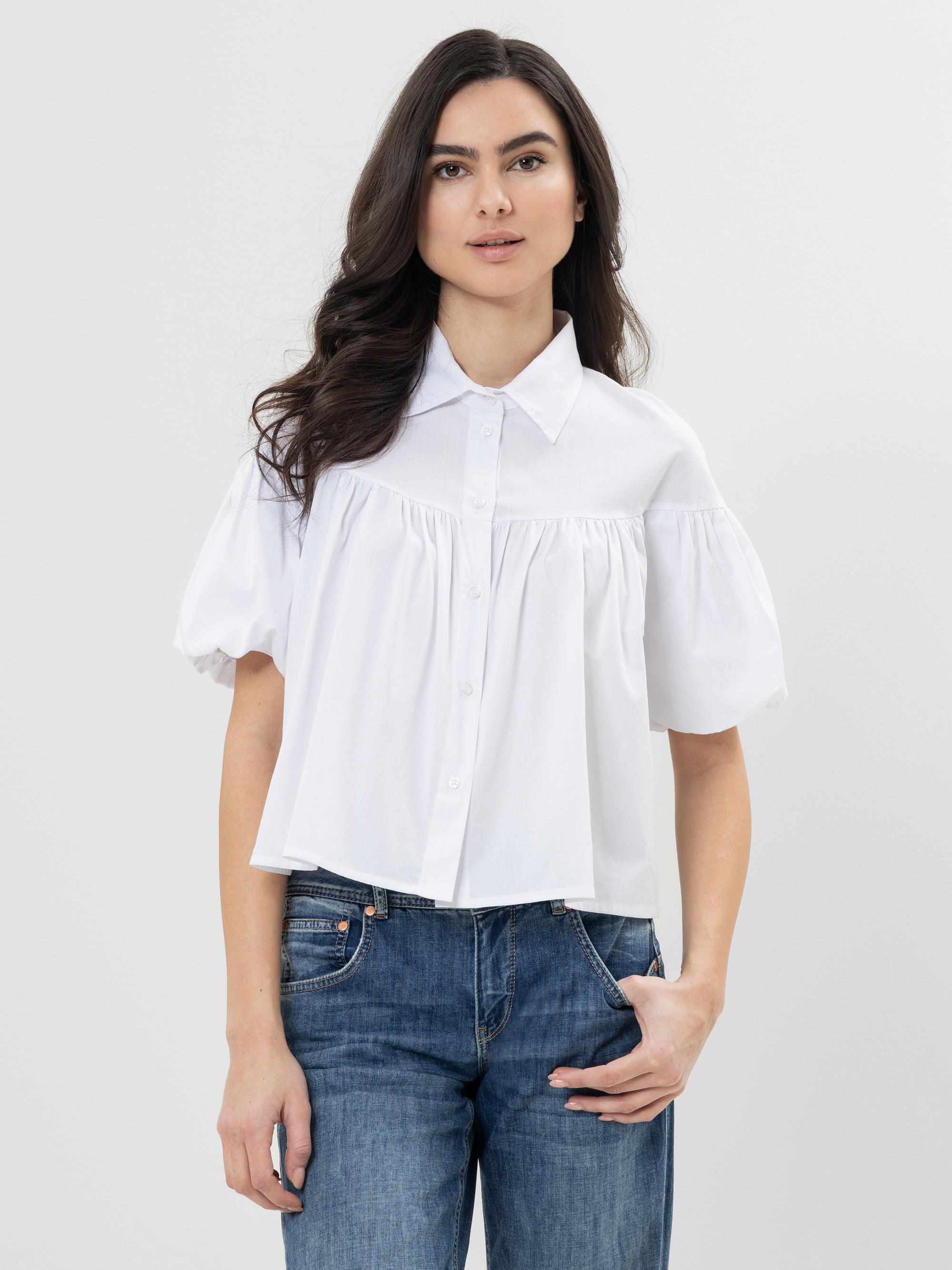 Eine Frau mit langen dunklen Haaren trägt eine locker sitzende weiße Button-up-Bluse mit kurzen Puffärmeln und blaue Jeans und steht vor einem schlichten hellen Hintergrund.