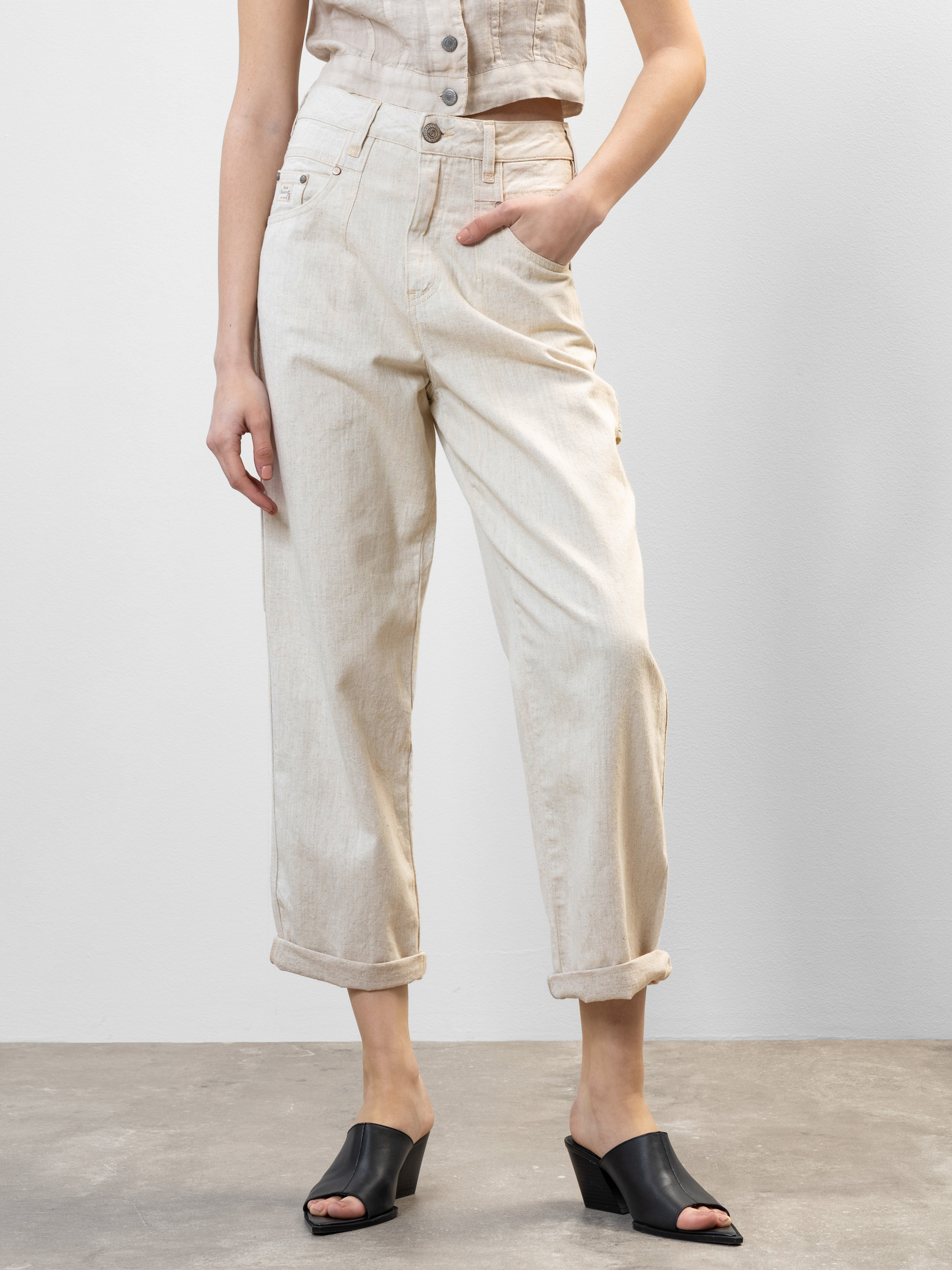 Eine Person steht auf einem grauen Boden und trägt eine hellbeige, hochgekrempelte Jeans, ein dazu passendes ärmelloses Oberteil und schwarze Pantoletten mit offenen Zehen. Eine Hand steckt in der Tasche, das Gesicht ist nicht zu sehen.