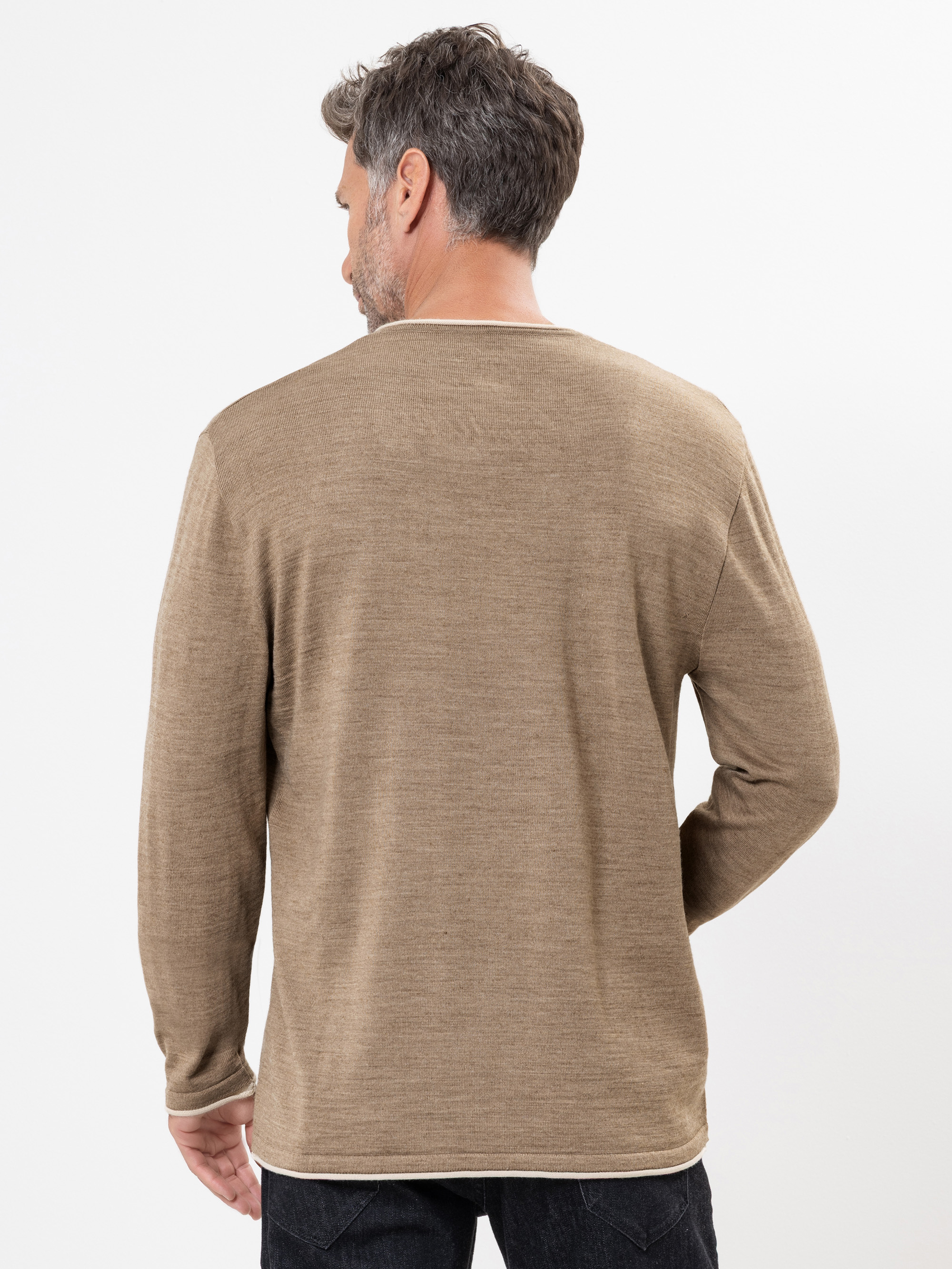 Ein Mann mit kurzen braunen Haaren, von hinten gesehen, trägt ein langärmeliges, hellbraunes Hemd und schwarze Jeans. Er steht vor einem schlichten weißen Hintergrund.